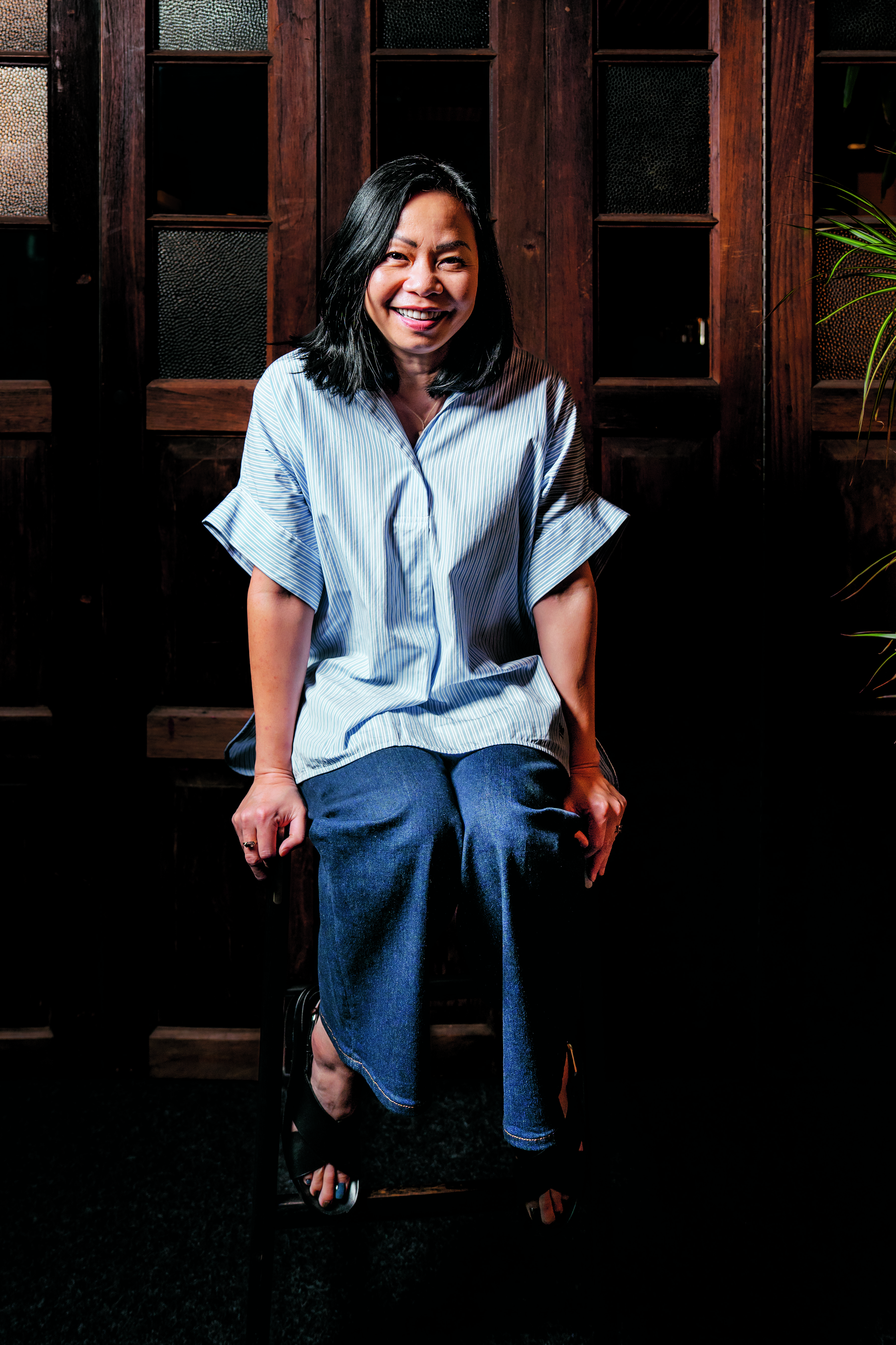 A smiling woman with black hair sits on a chair, wearing a striped blue blouse and dark jeans, in front of wooden doors.