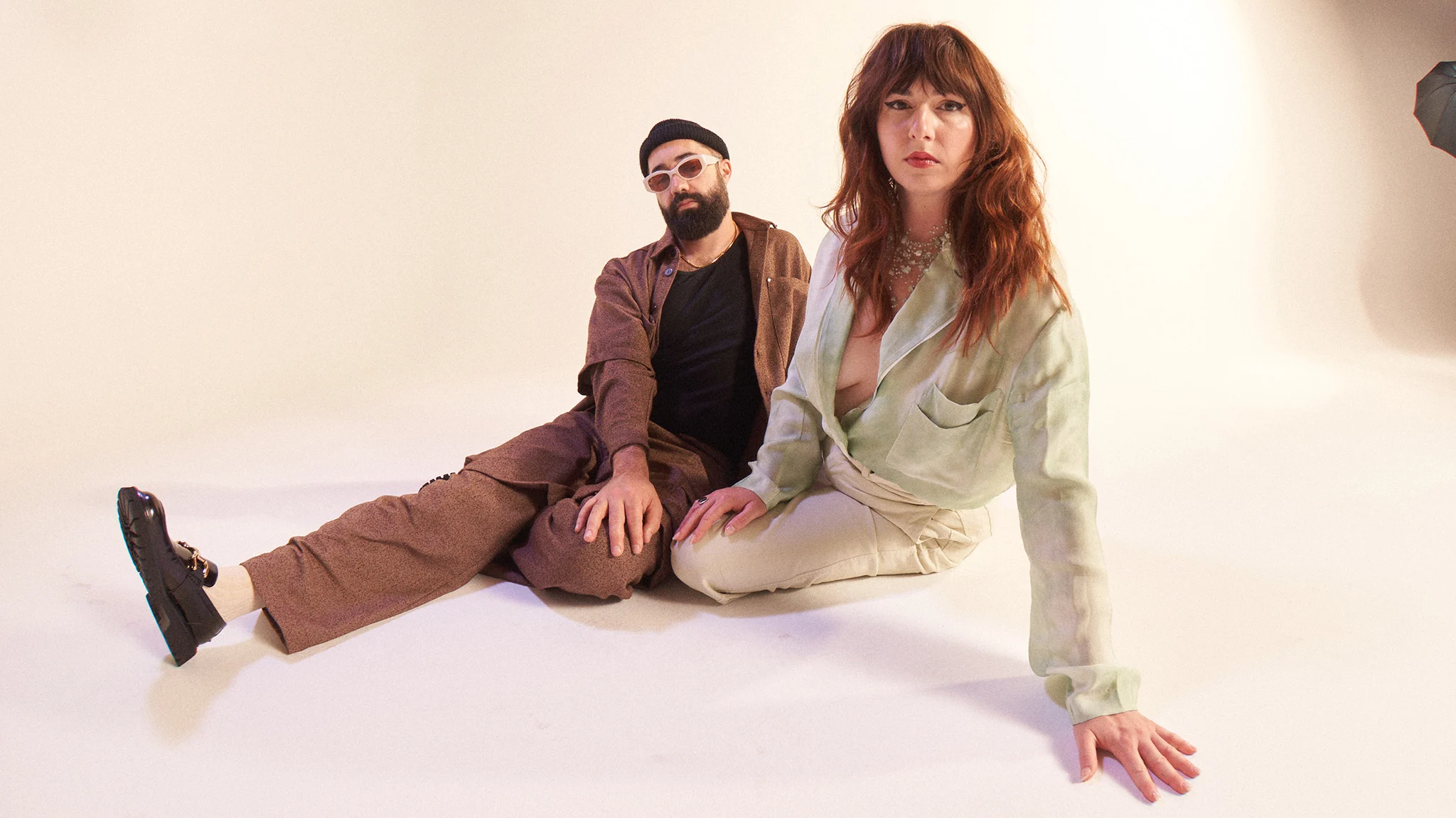 Two people sit on a white studio floor; a bearded man in brown and a red-haired woman in a sage green top.