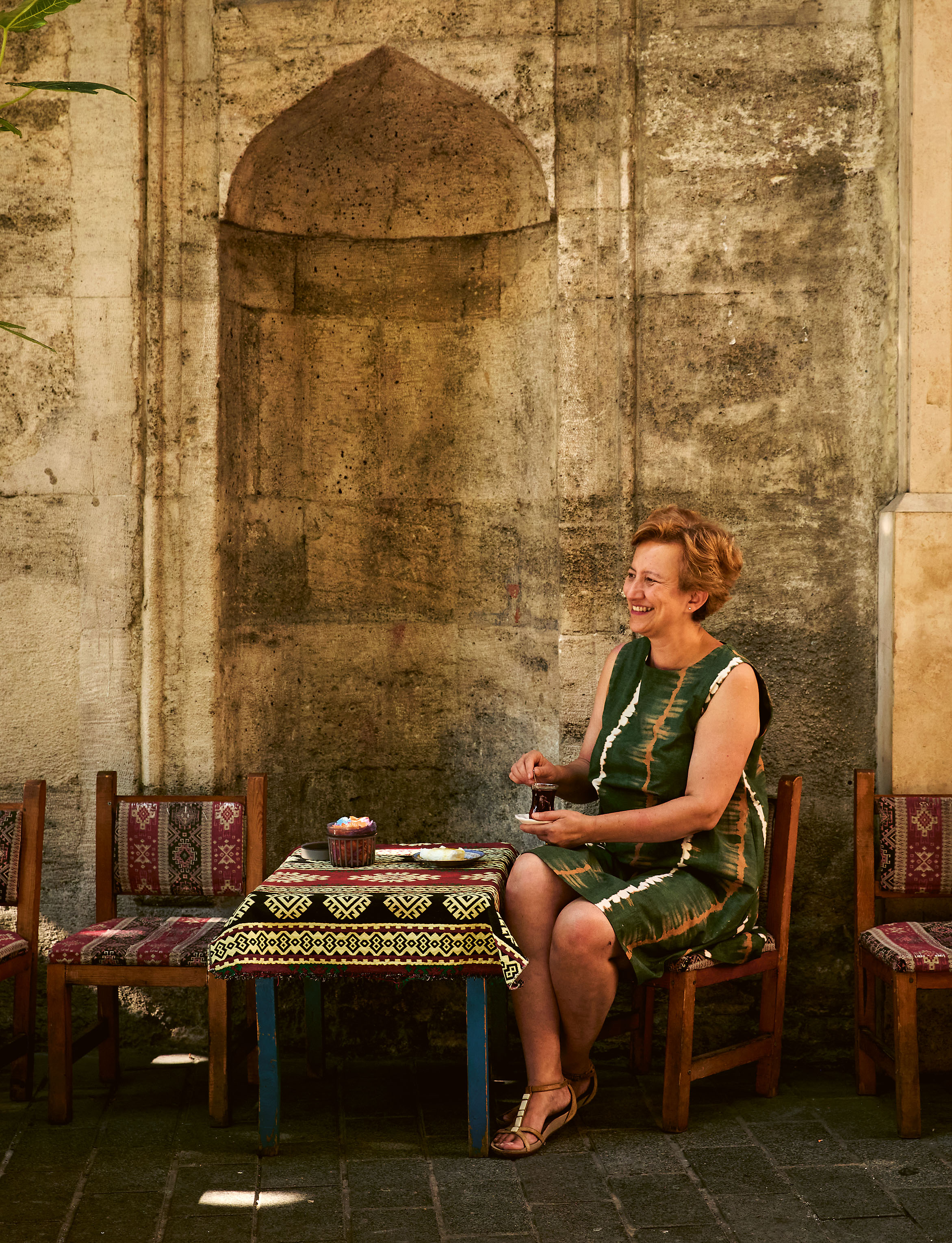 A smiling woman in a green dress sips tea at a patterned table beside an ancient stone wall with an arched niche.