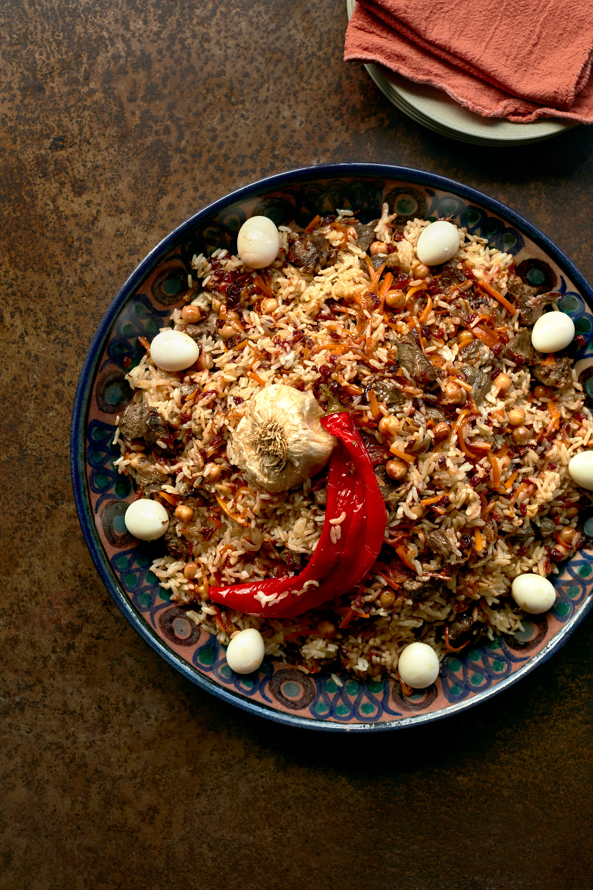 Traditional rice dish with meat, chickpeas, and quail eggs in a decorative blue plate, topped with red pepper and garlic.