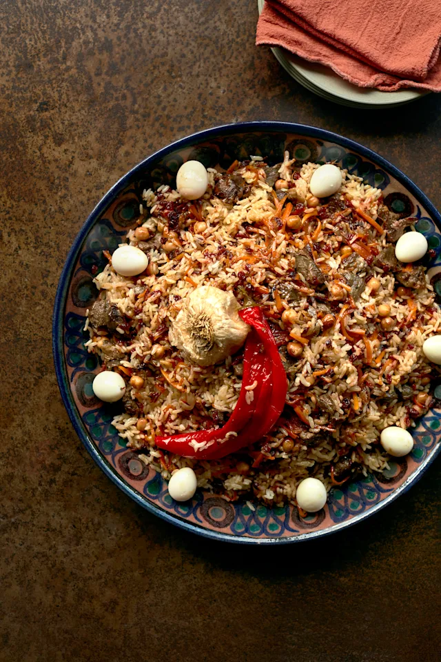 Traditional rice dish with meat, chickpeas, and quail eggs in a decorative blue plate, topped with red pepper and garlic.