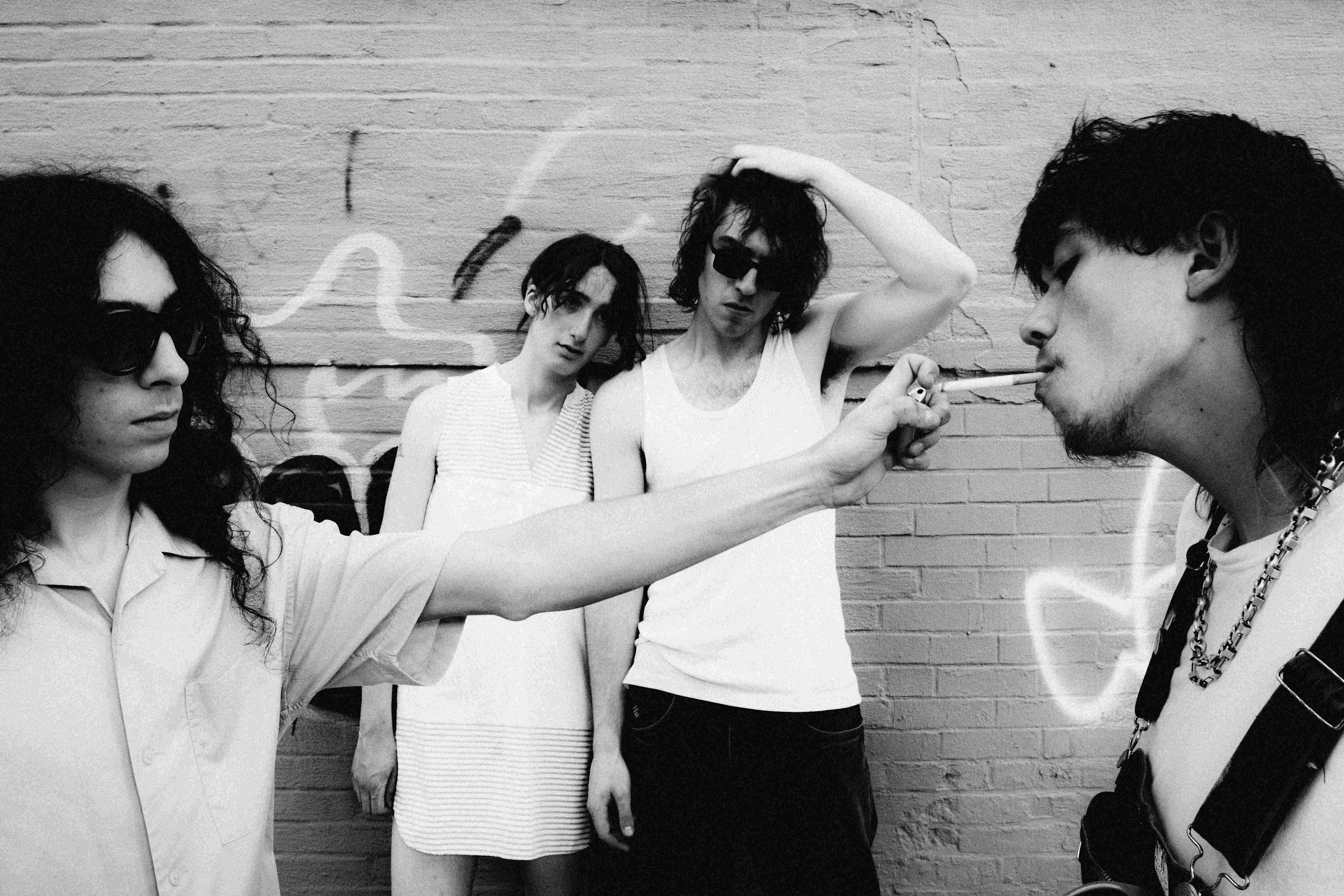 Four young musicians in black and white photo against brick wall, one lighting a cigarette for another, edgy rock band aesthetic.