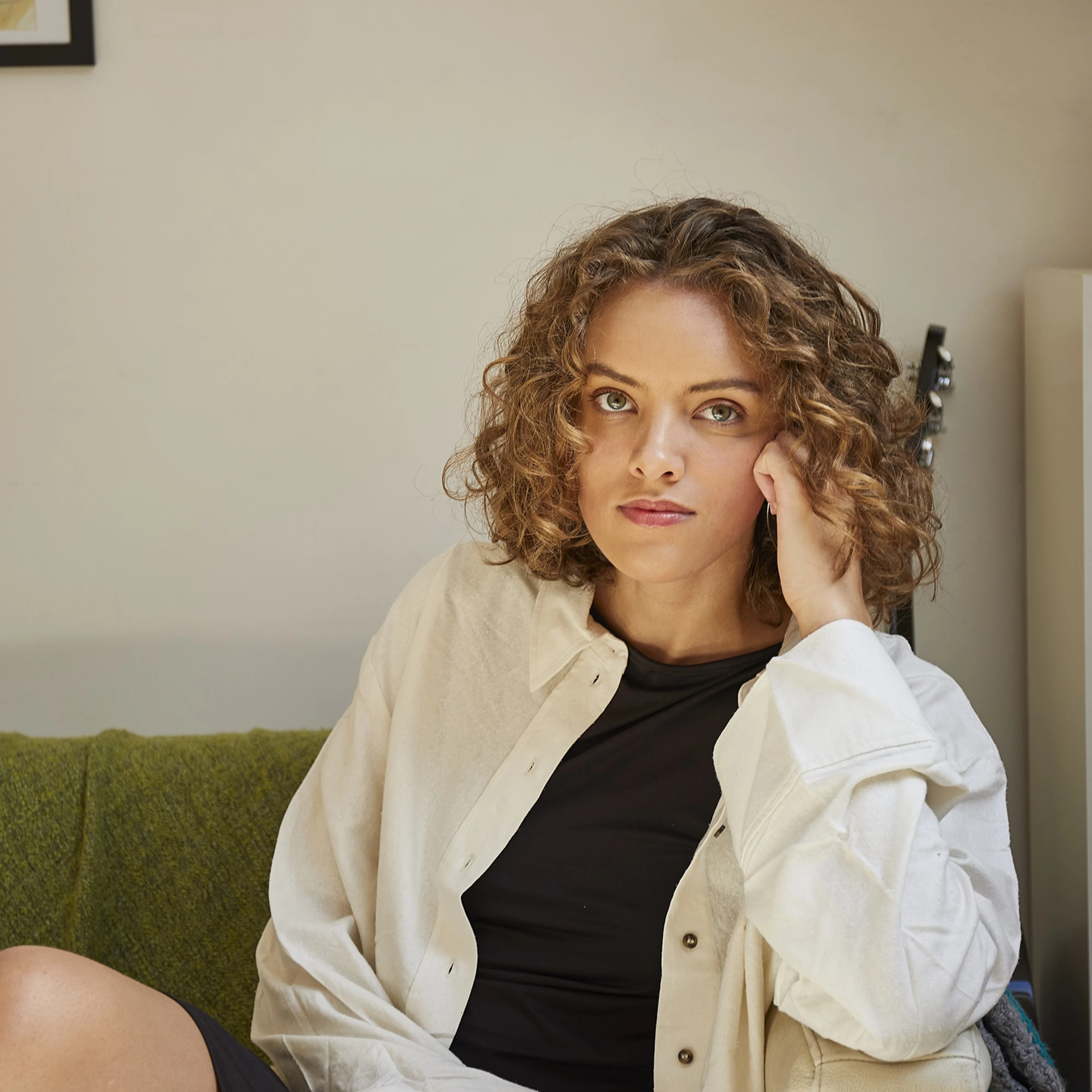 Person with curly hair wearing black dress and white overshirt sitting on green couch, framed artwork on wall above.
