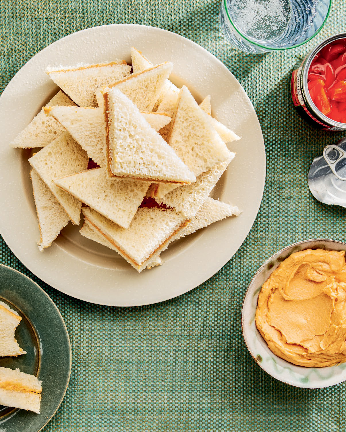 Triangular sandwich pieces on white plate with hummus dip, roasted peppers, and water glass on green tablecloth.