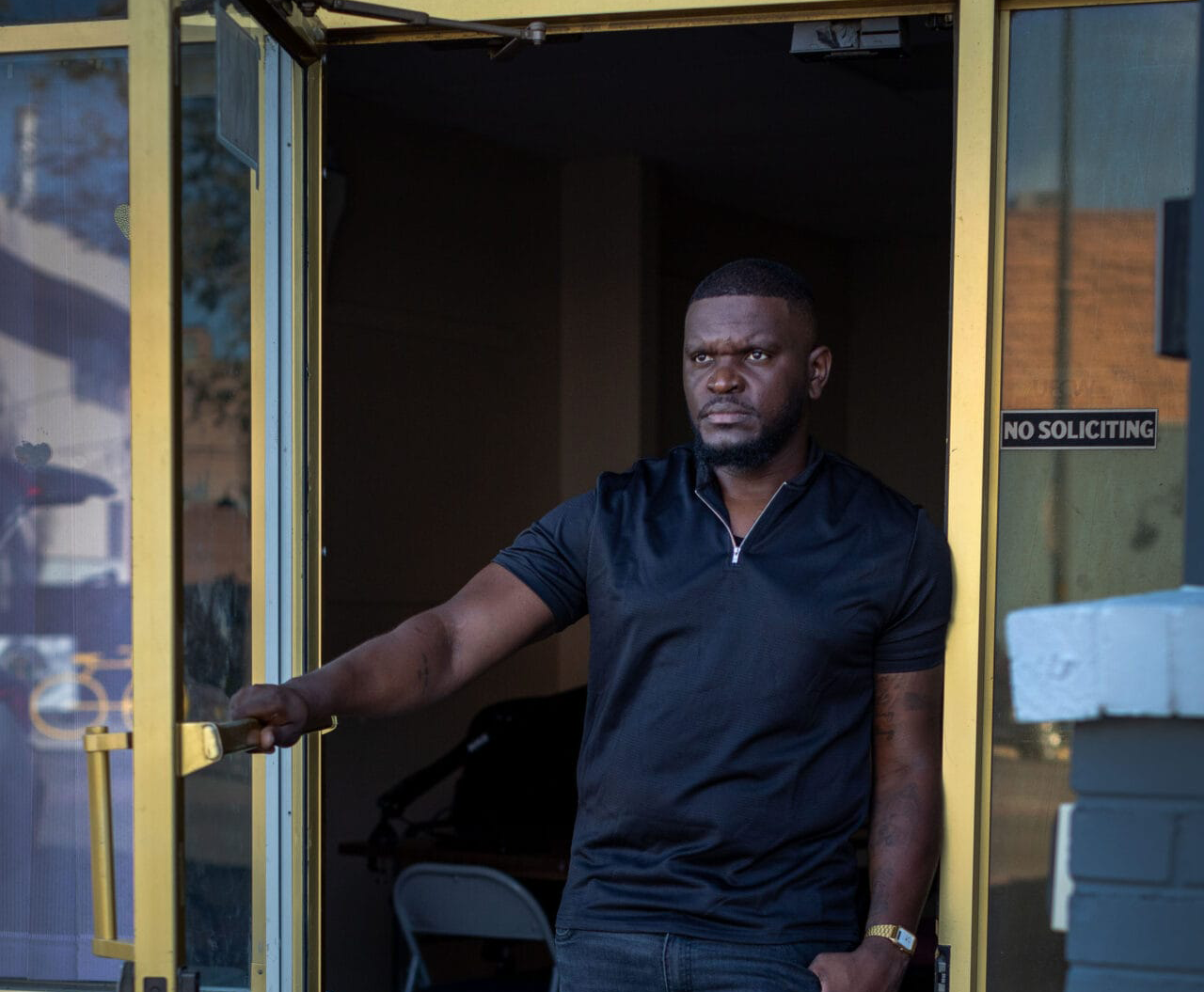 A man in a black shirt stands in a yellow-framed glass doorway, holding the door open, with a "No Soliciting" sign nearby.