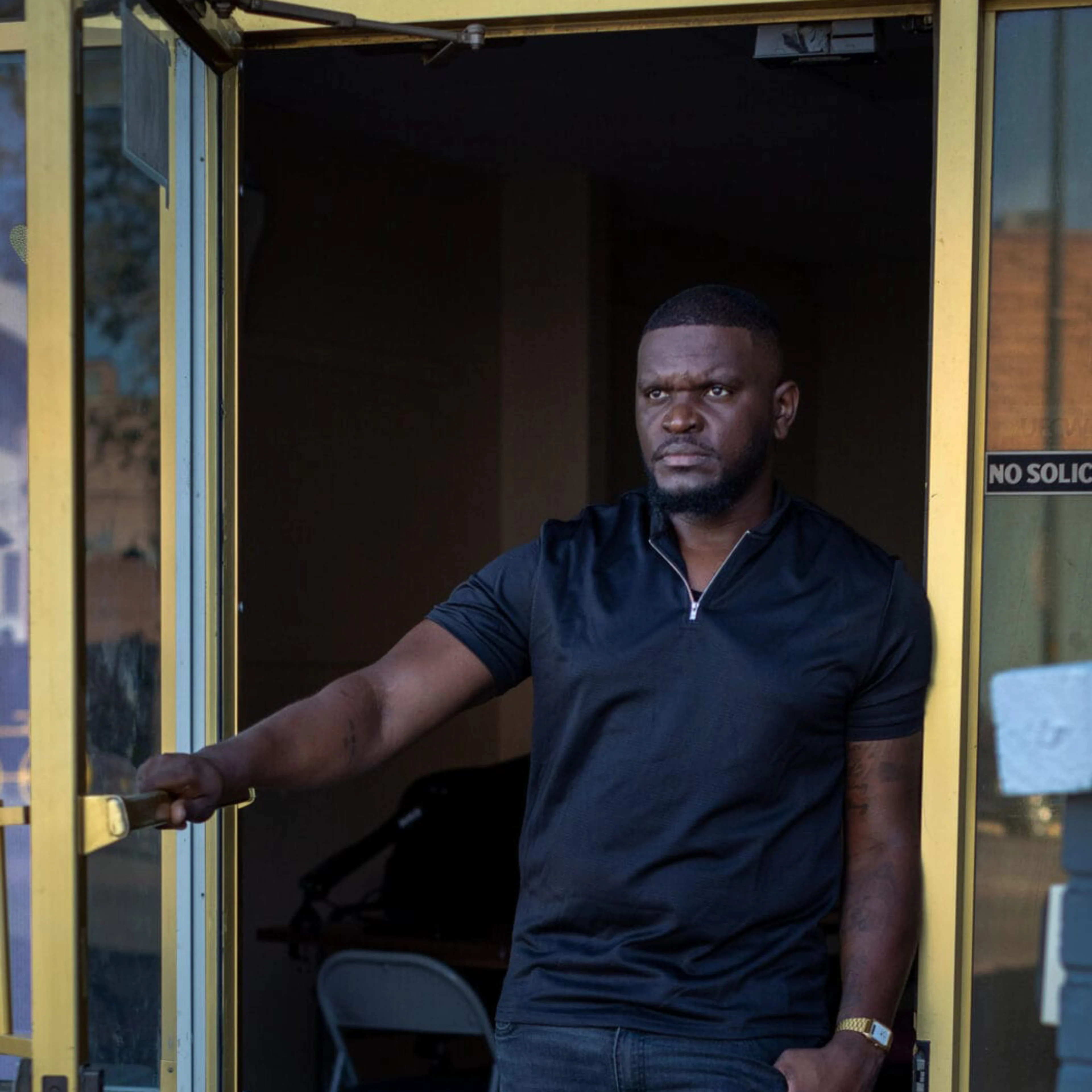A man in a black shirt stands in a yellow-framed glass doorway, holding the door open, with a "No Soliciting" sign nearby.