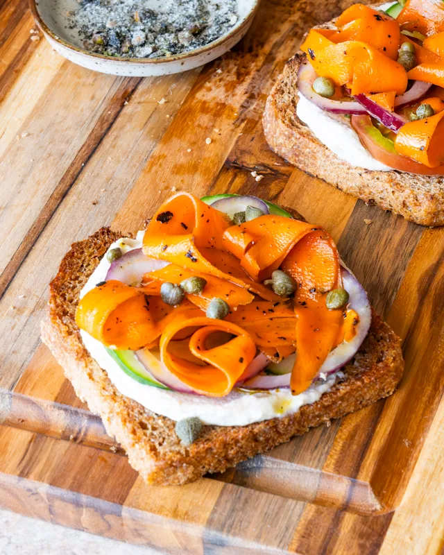 Two open-faced toasts on a wooden board topped with cream cheese, orange carrot ribbons, capers, and red onion.