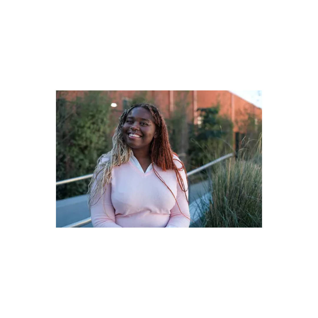 Person with long braided hair smiling outdoors, wearing a light pink top against a backdrop of greenery and brick wall.