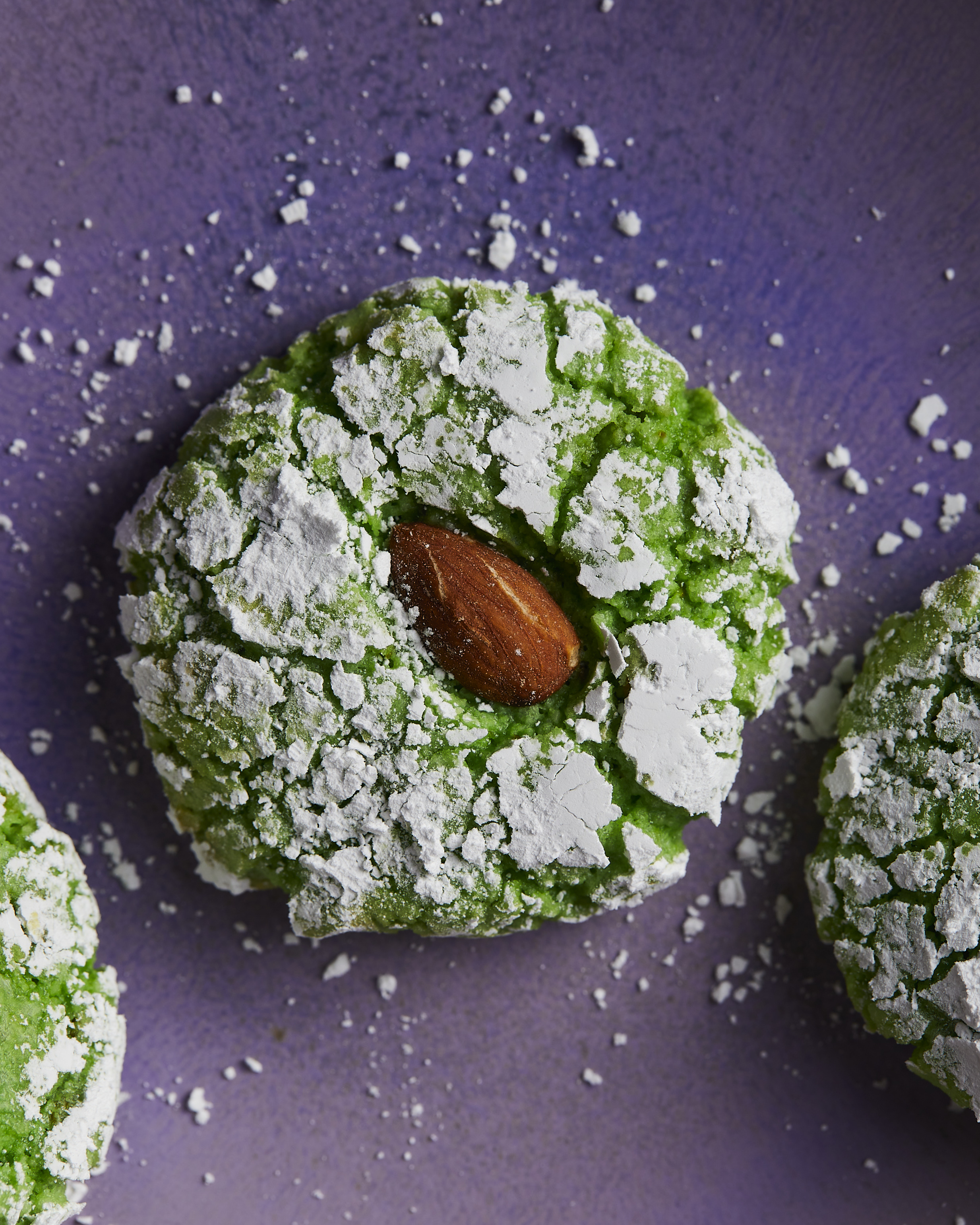Green crinkle cookie dusted with powdered sugar and topped with an almond on purple background.