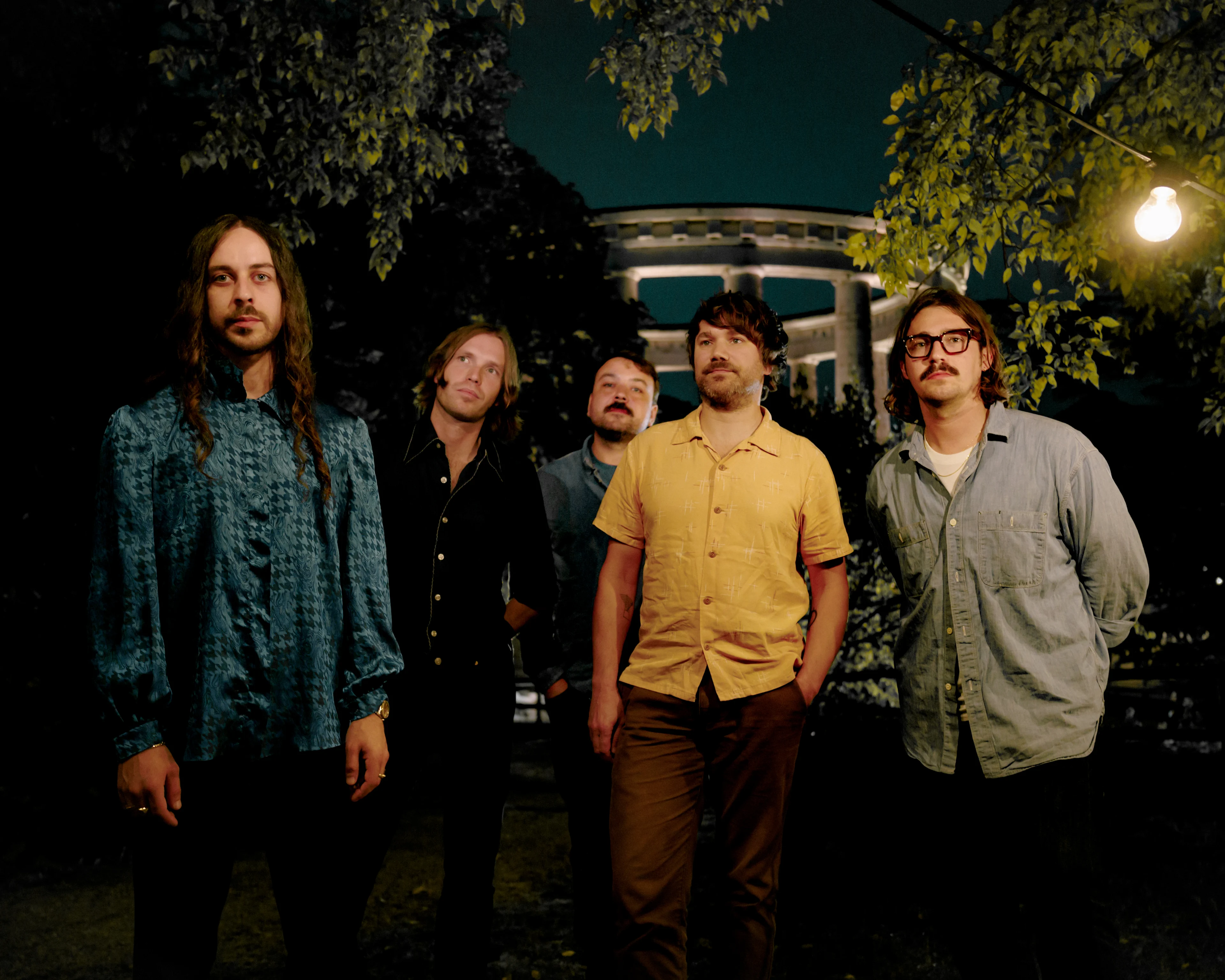 Five men standing outdoors at night near a lit gazebo and trees, dressed in casual shirts in blue, black, yellow, and gray.