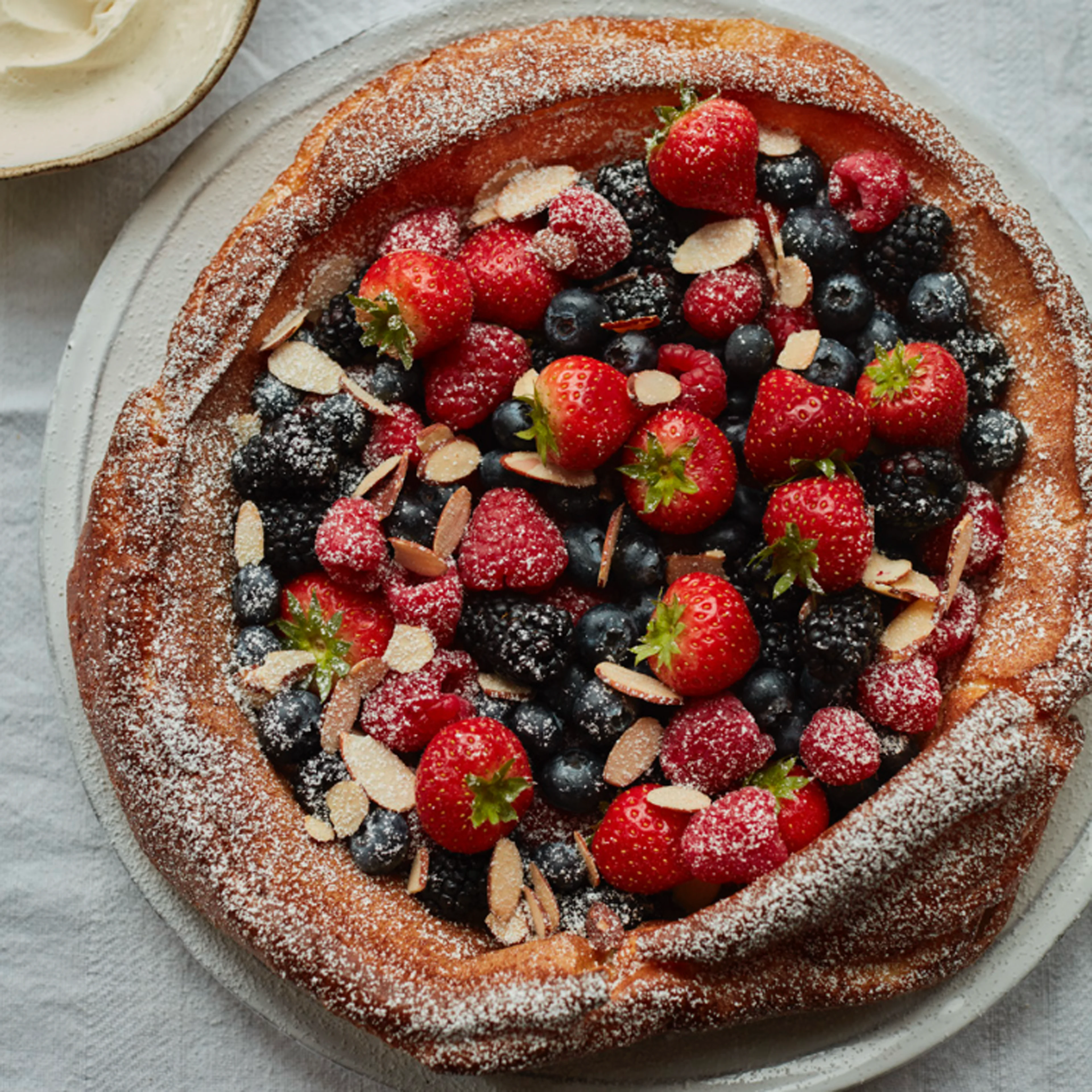 A golden baked tart topped with fresh strawberries, blueberries, blackberries, raspberries, sliced almonds, and powdered sugar.