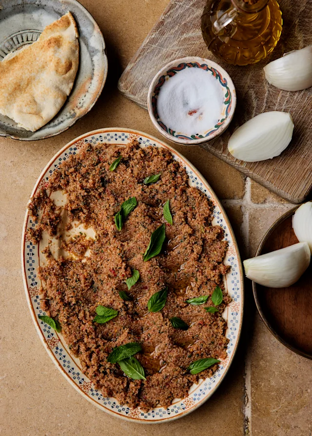 A decorative oval plate of kibbeh garnished with fresh mint, alongside pita bread, salt, olive oil, and onions.