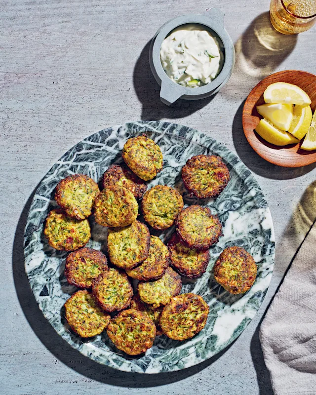 Golden-brown zucchini fritters on a marble plate, served with tzatziki dip and lemon wedges on the side.