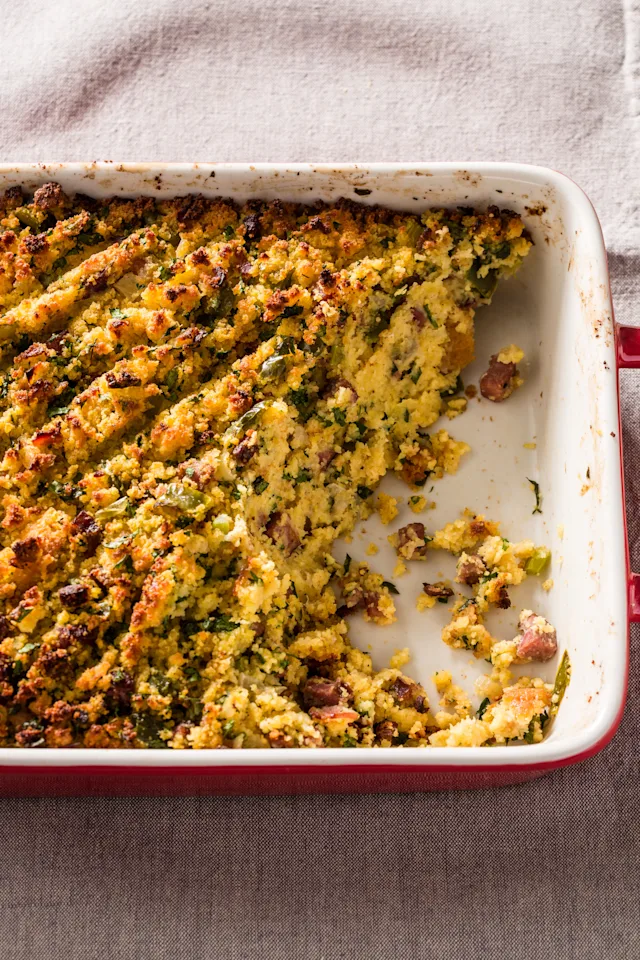 Golden cornbread stuffing in a white baking dish with red trim, showing herbs and sausage pieces mixed throughout.
