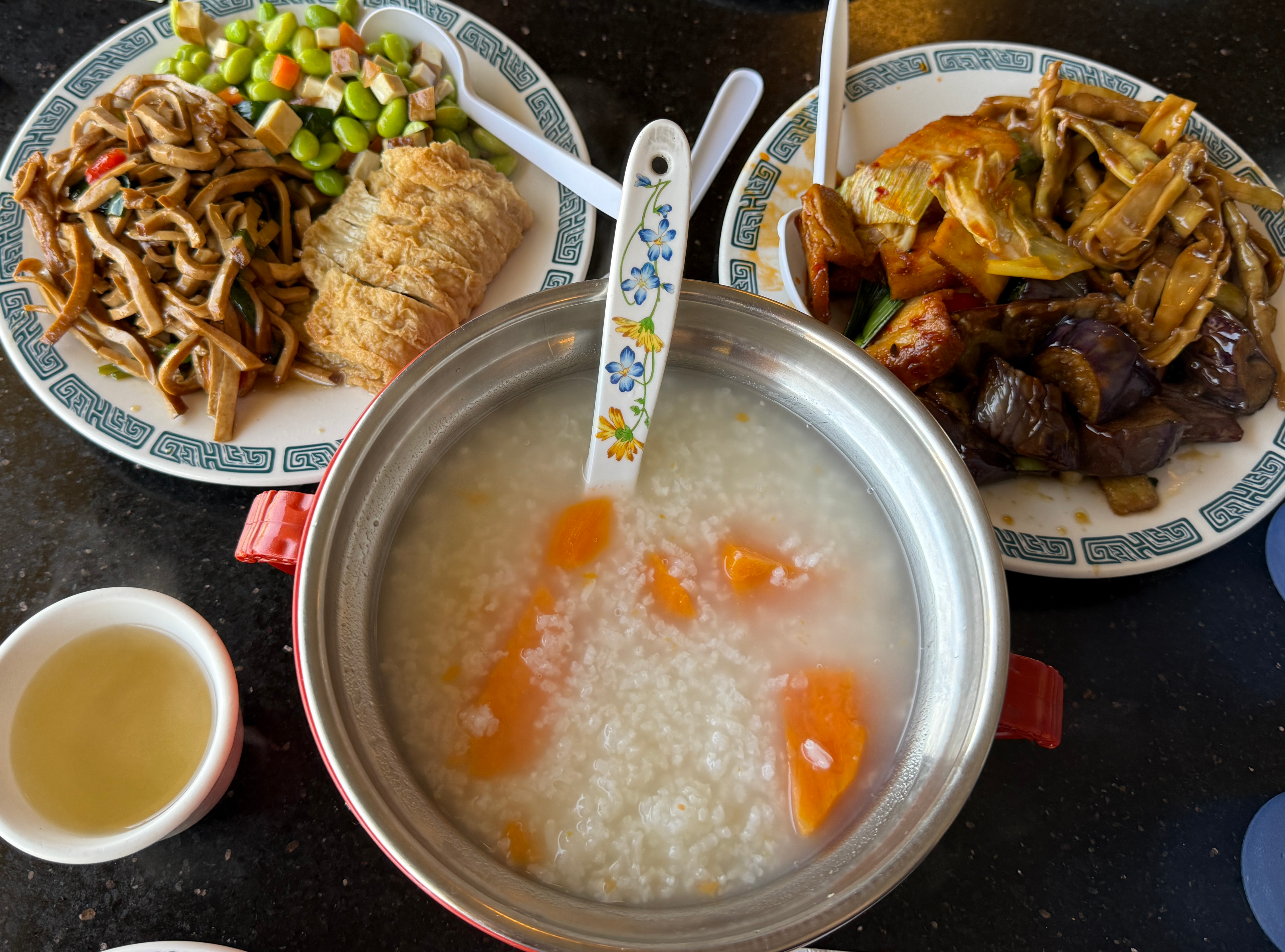 A spread of Chinese dishes including a pot of congee with orange pieces, stir-fried vegetables, tofu, and a cup of broth.