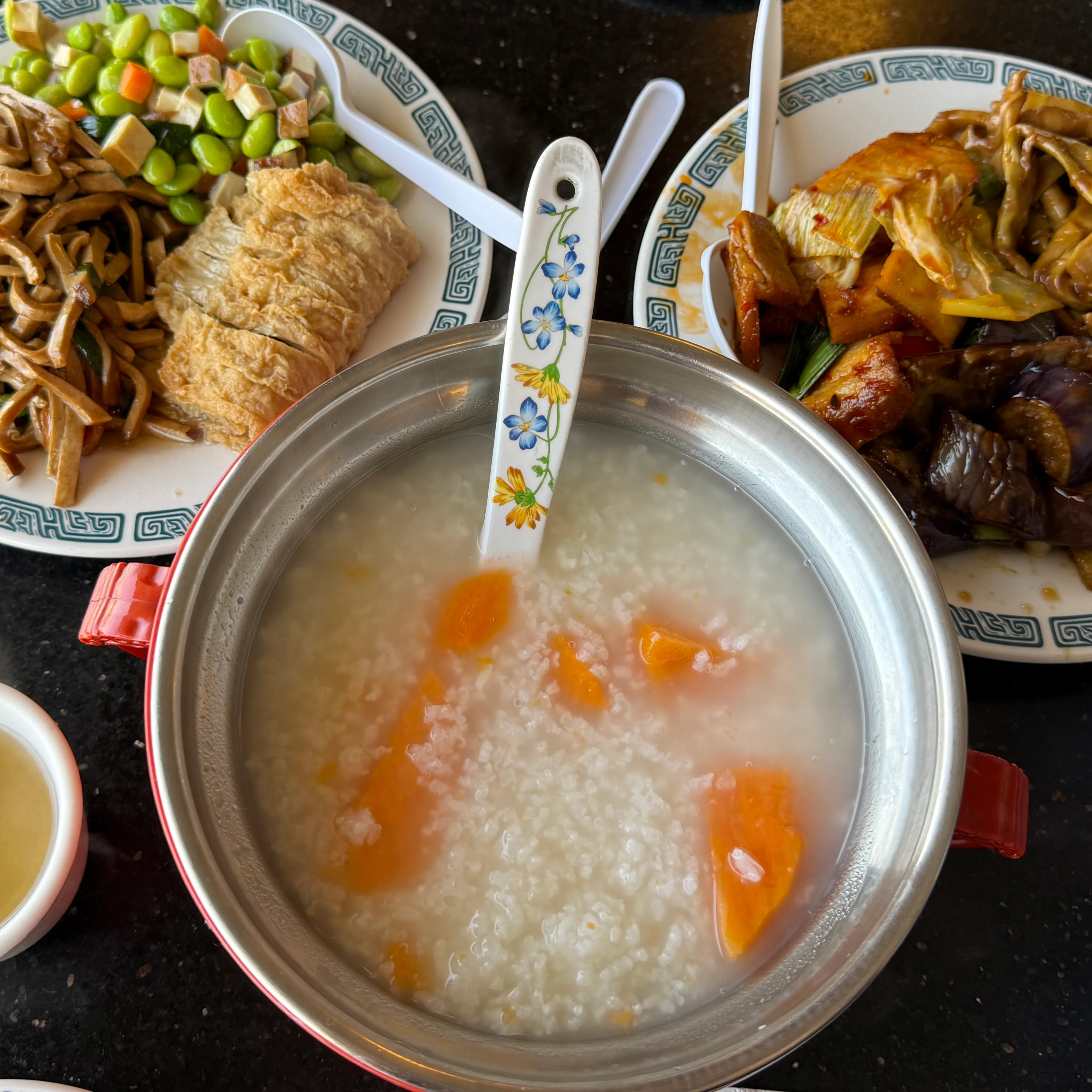 A spread of Chinese dishes including a pot of congee with orange pieces, stir-fried vegetables, tofu, and a cup of broth.
