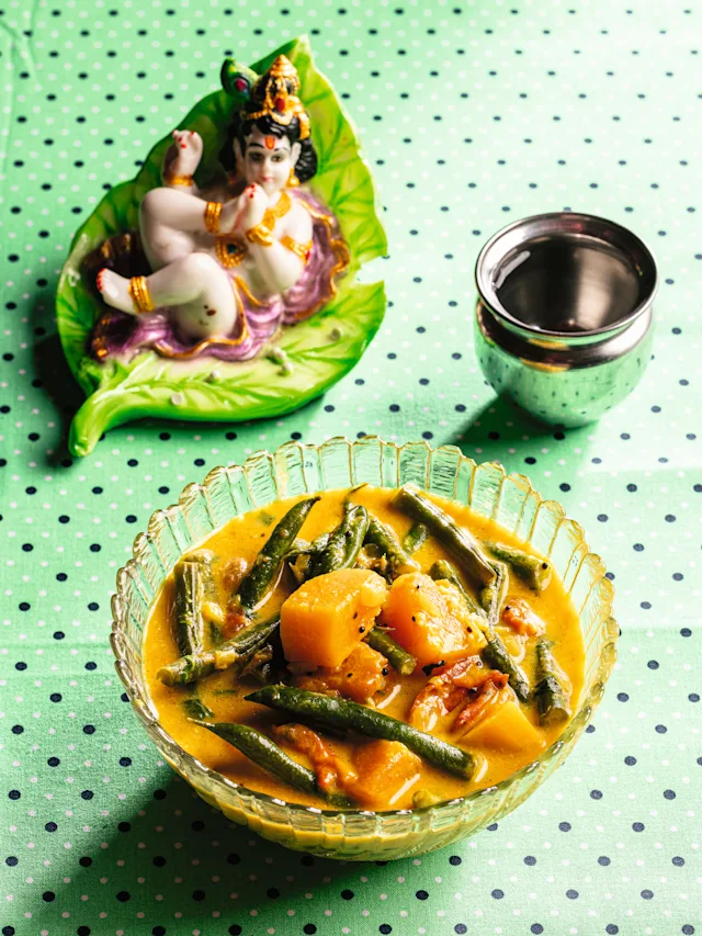 A glass bowl of yellow vegetable curry with green beans on a polka-dot cloth, beside a Hindu deity figurine and steel cup.