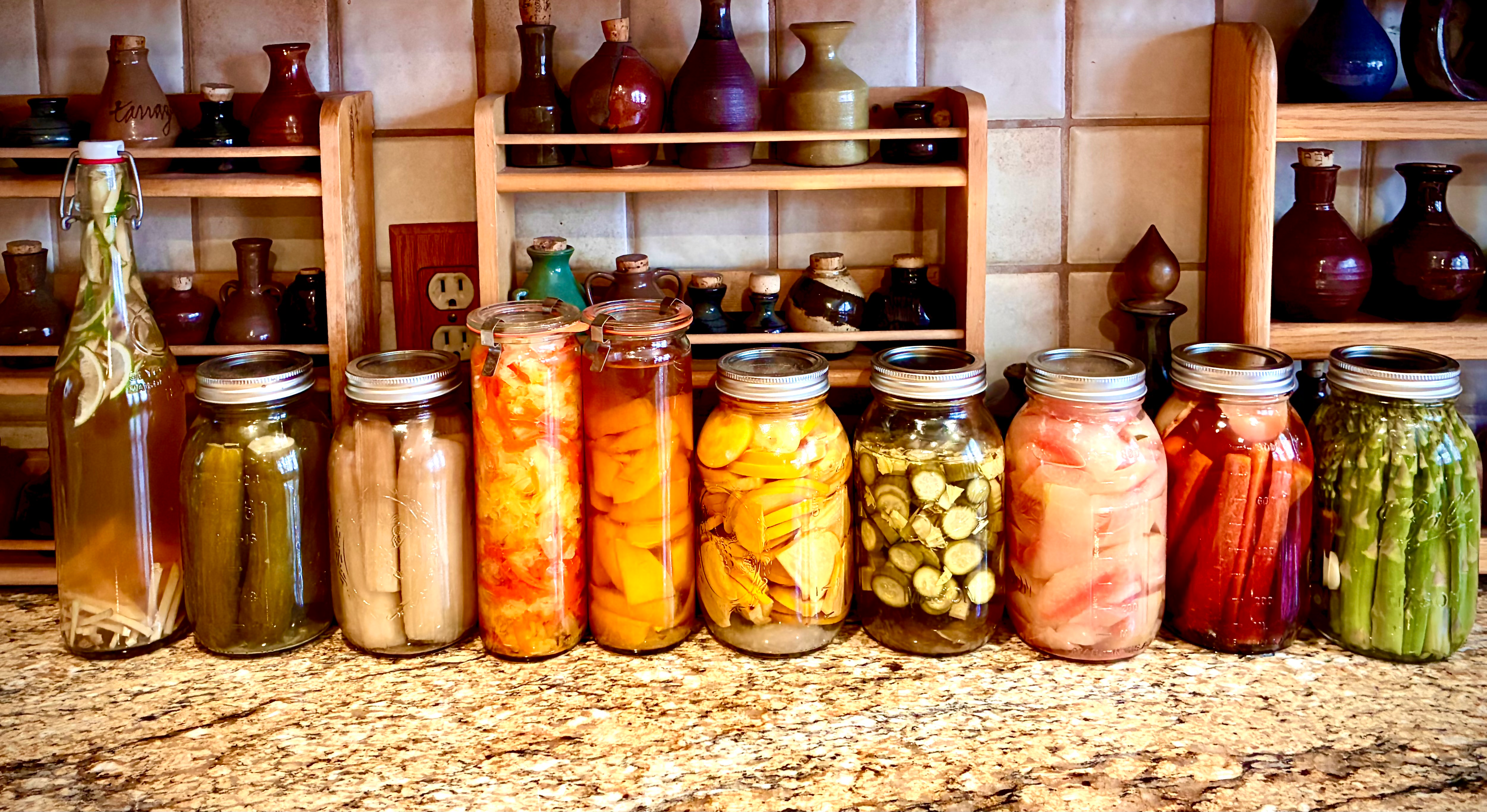 A row of colorful home-canned vegetable jars on a granite counter, with ceramic pottery on wooden shelves behind them.