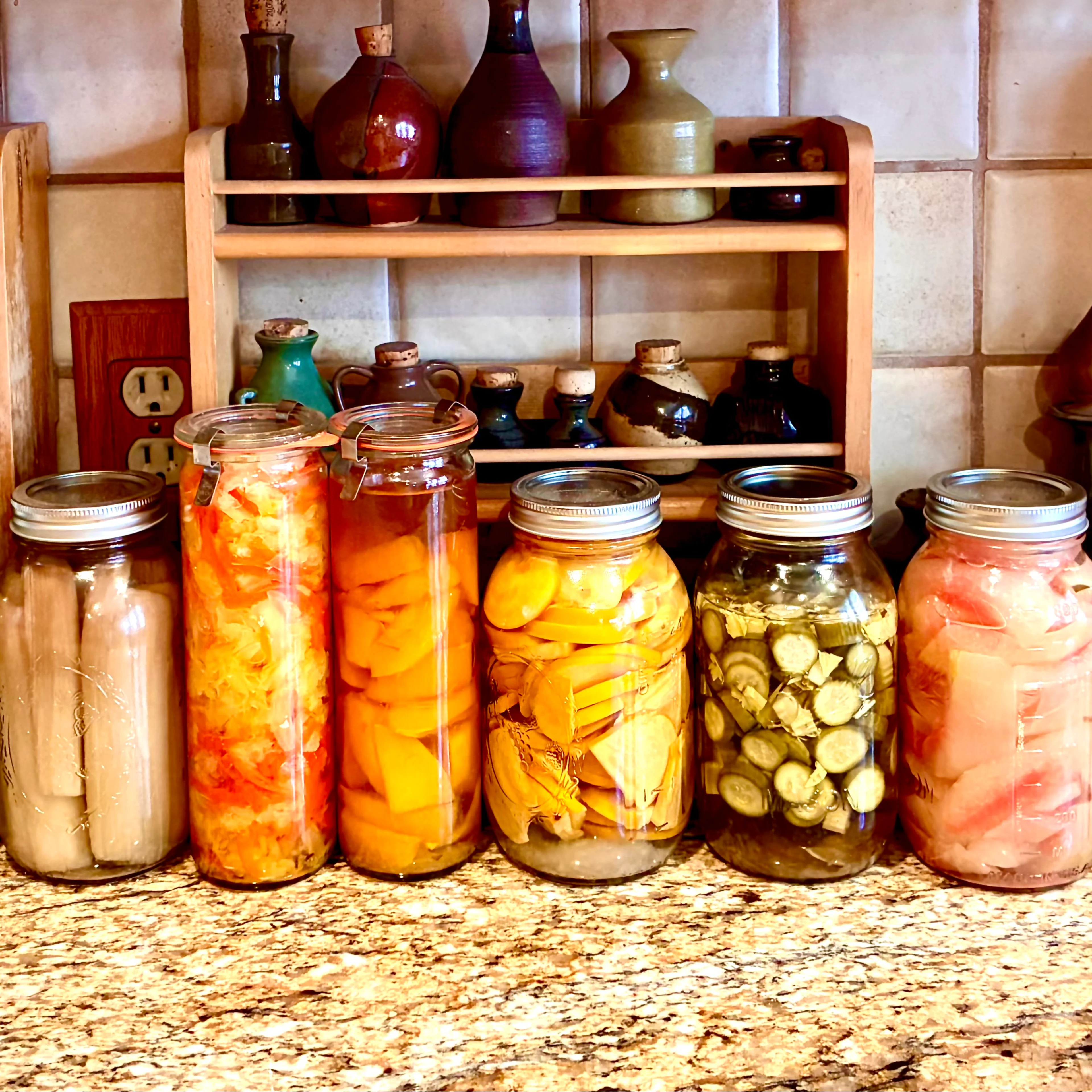 A row of colorful home-canned vegetable jars on a granite counter, with ceramic pottery on wooden shelves behind them.