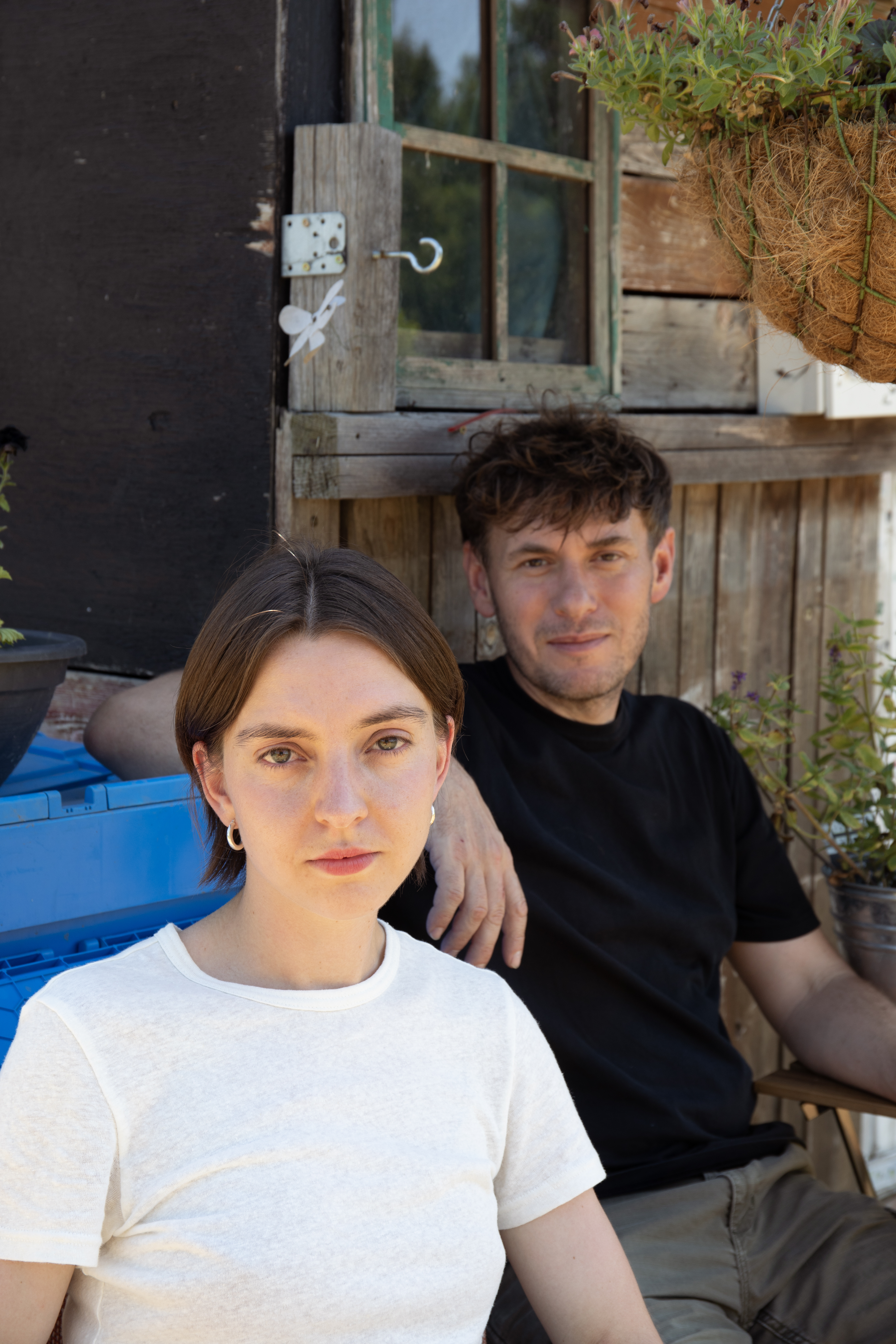 Two people sit outdoors near a rustic wooden shed with hanging plants. A woman in white and a man in black look at the camera.