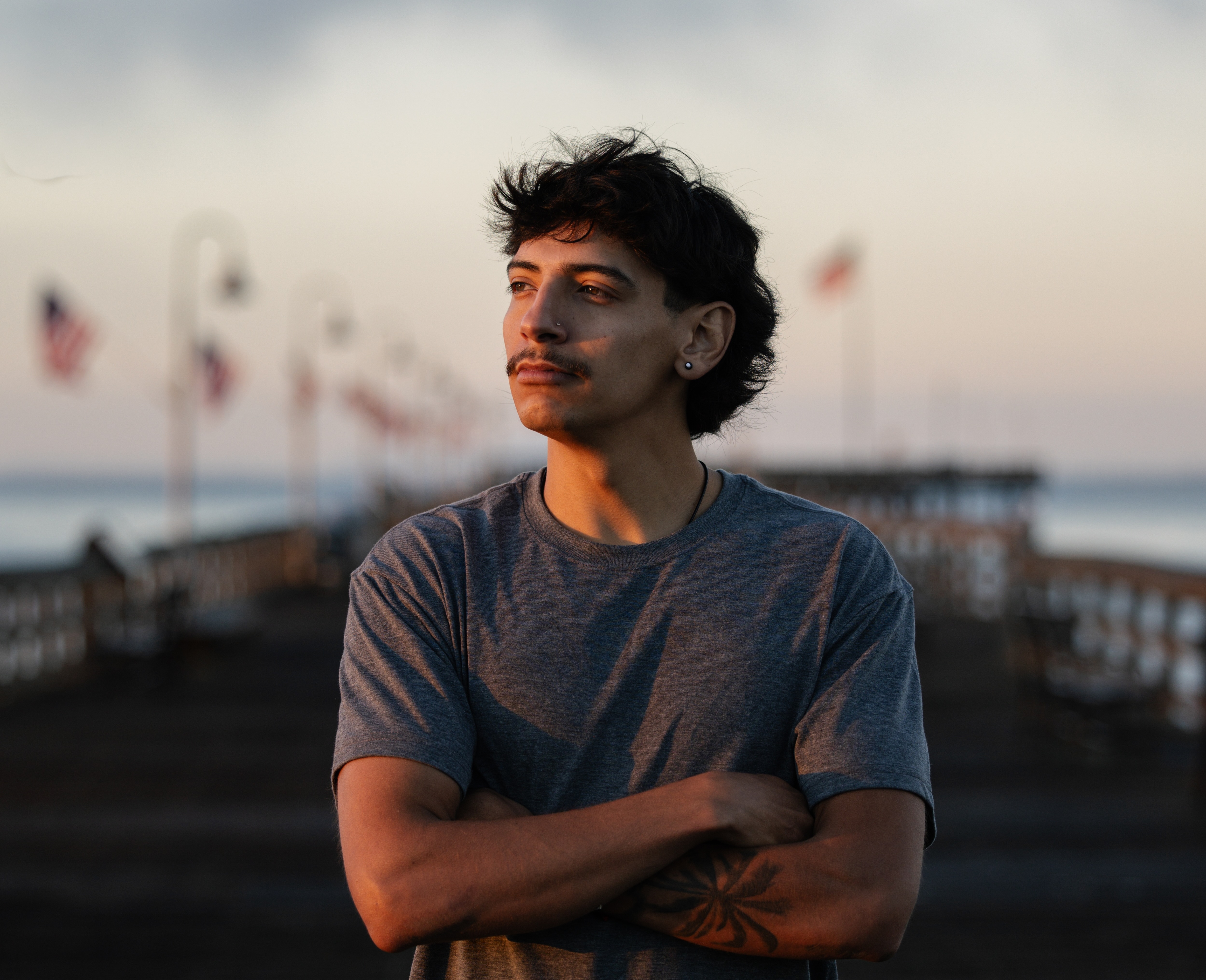 A Latino man with dark, curly hair poses on a wooden boardwalk with American flags in the background. 