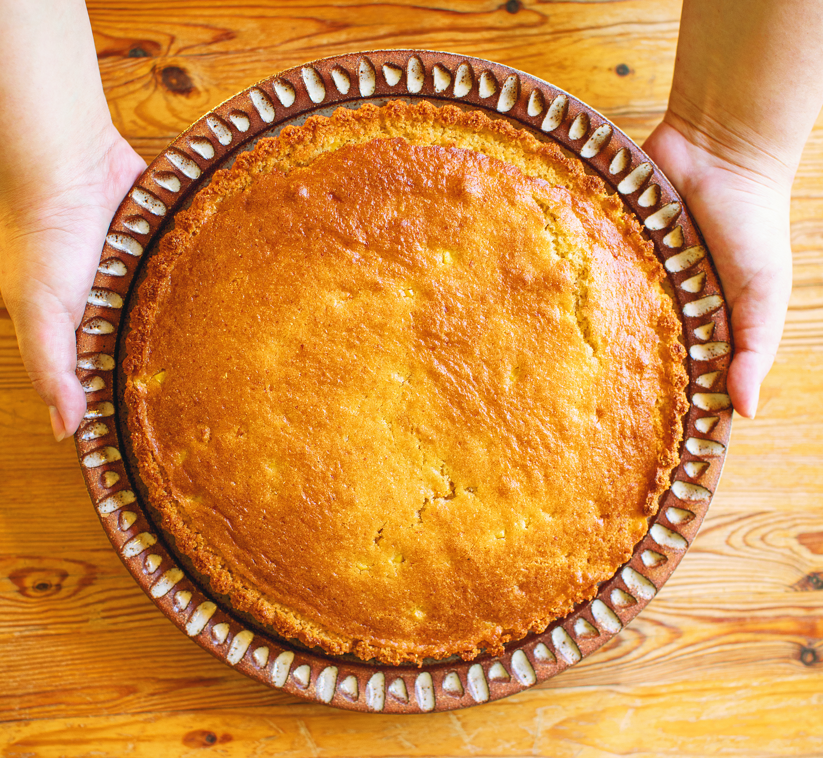 Hands holding a freshly baked golden pie or tart in a decorative ceramic dish on a wooden table.