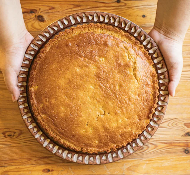 Hands holding a freshly baked golden pie or tart in a decorative ceramic dish on a wooden table.
