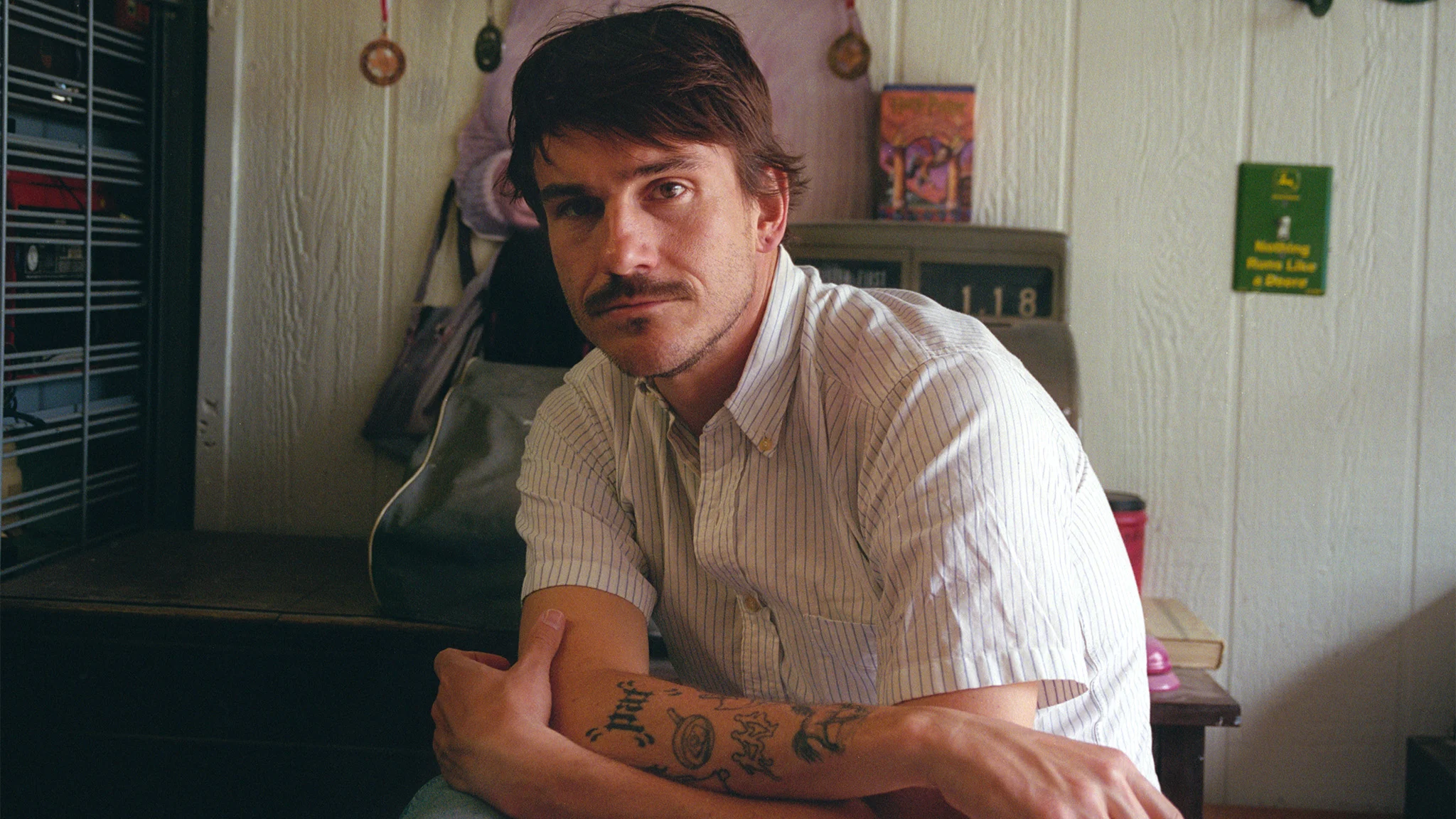 A young man with a mustache and arm tattoos sits with arms crossed, wearing a white striped shirt in a cluttered room.