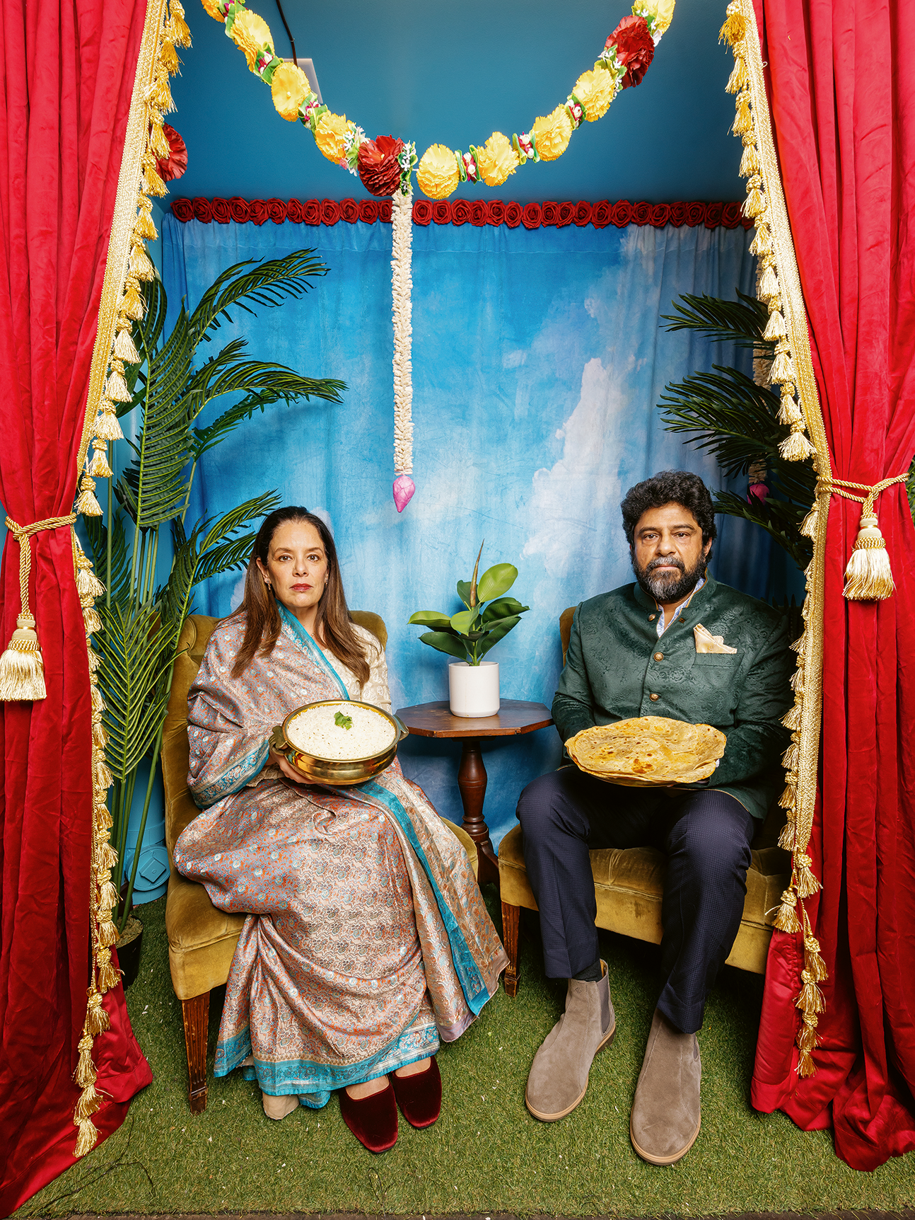 Two people in traditional South Asian attire sit holding dishes of food, framed by red curtains and colorful floral garlands.