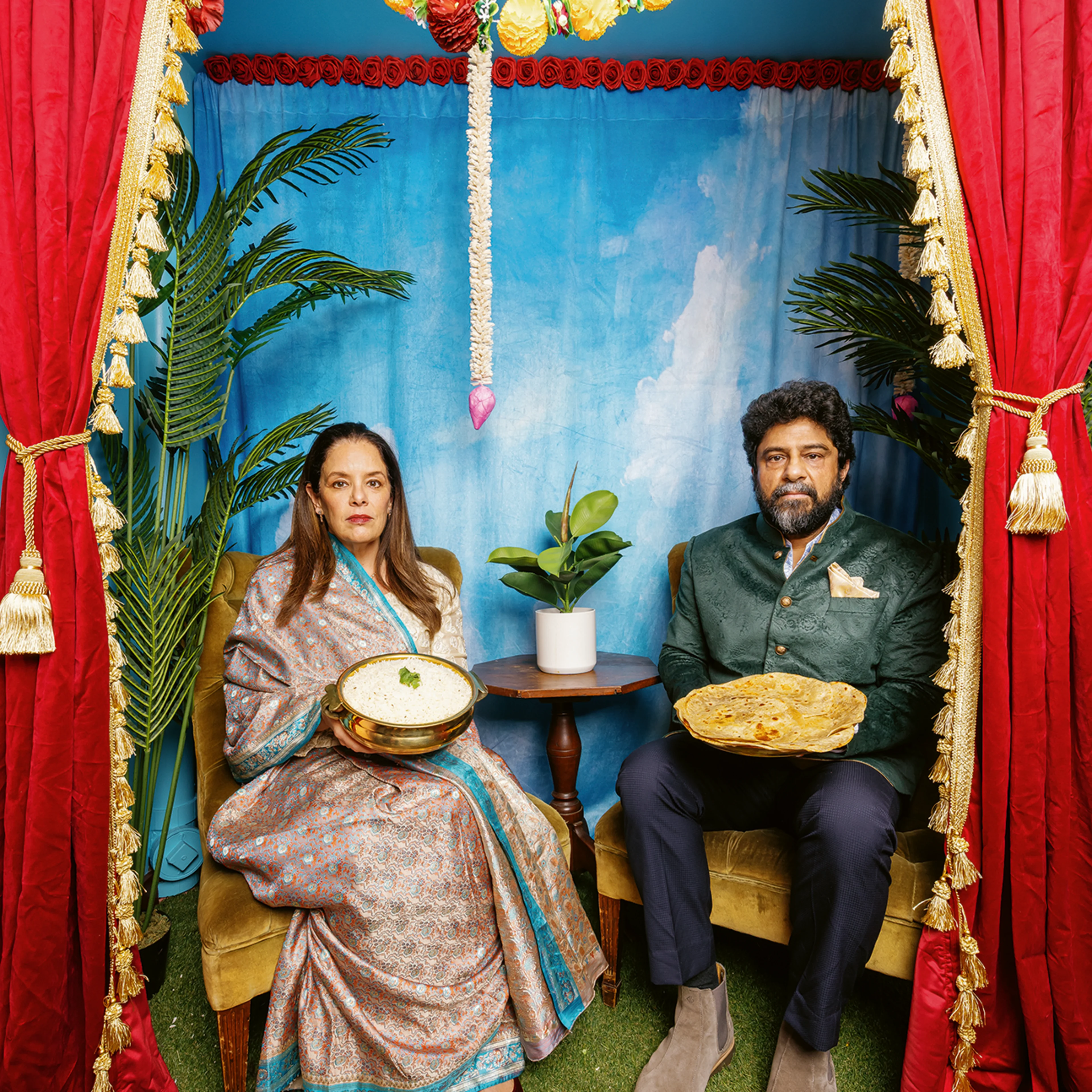 Two people in traditional South Asian attire sit holding dishes of food, framed by red curtains and colorful floral garlands.