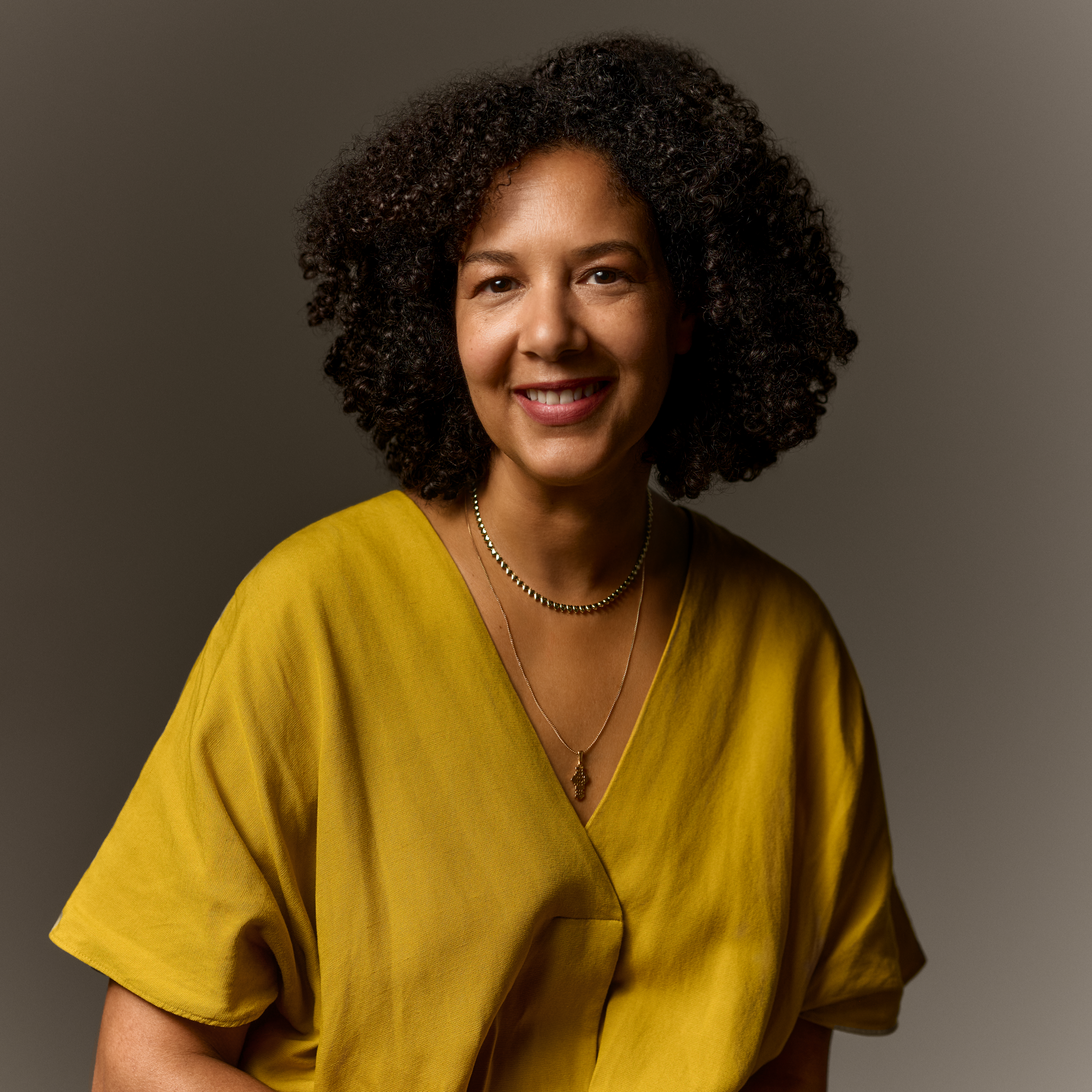 Woman in a ocre shirt with two necklaces and dark curly hair.