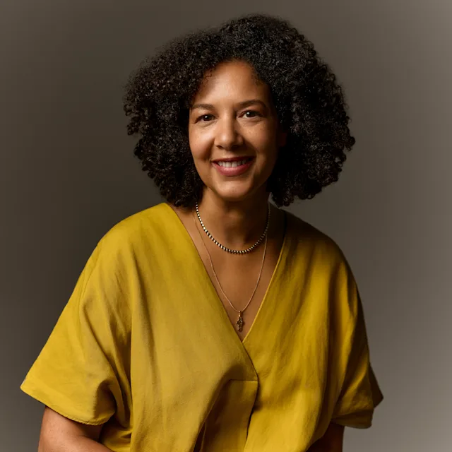 Woman in a ocre shirt with two necklaces and dark curly hair.