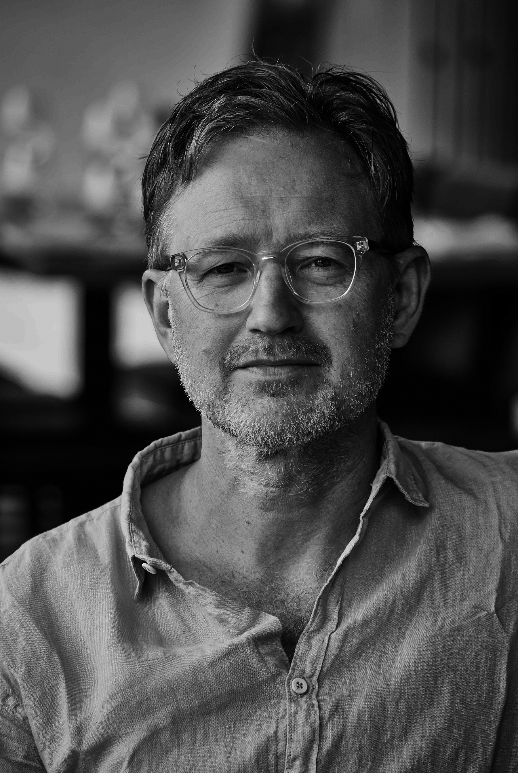 Black and white portrait of a middle-aged man with clear-framed glasses, stubble, and an open-collar linen shirt.