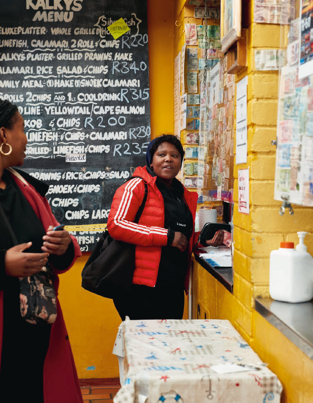 A woman in a red jacket stands at Kalky's seafood counter, smiling, with a chalkboard menu and yellow walls behind her.