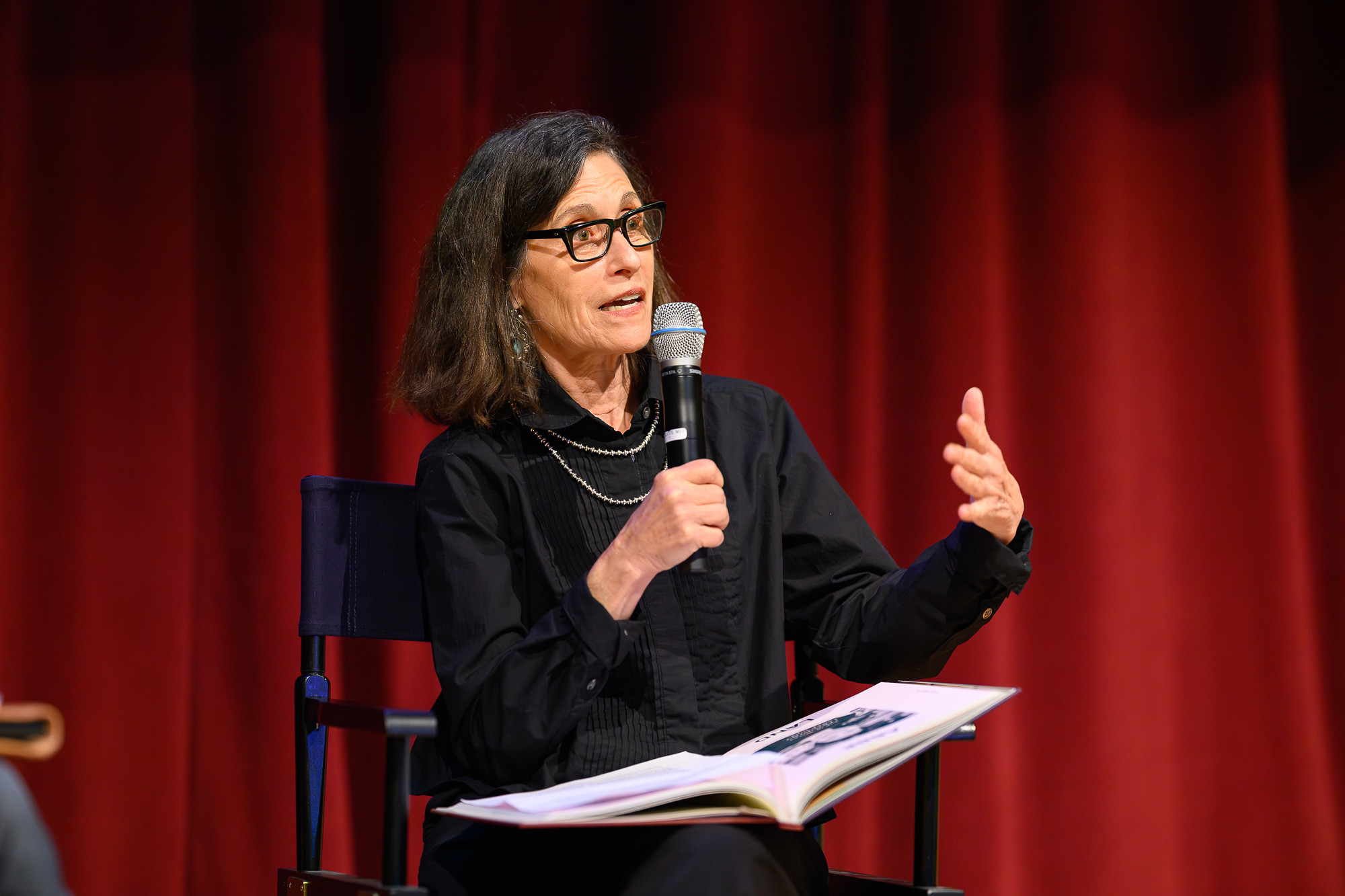 An woman with a microphone sits in a director's chair on a stage describing a book she wrote, which sits in her lap.