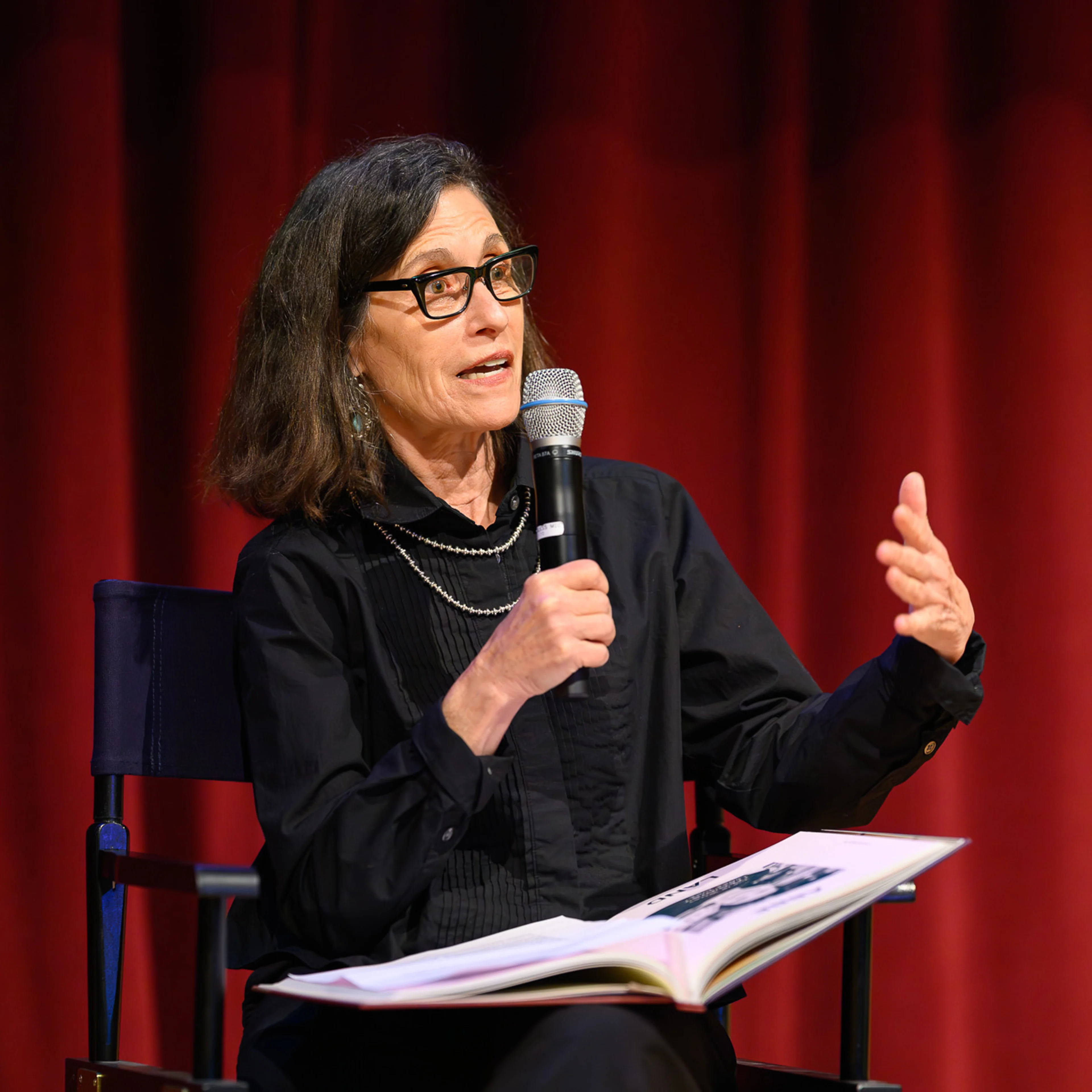 An woman with a microphone sits in a director's chair on a stage describing a book she wrote, which sits in her lap.