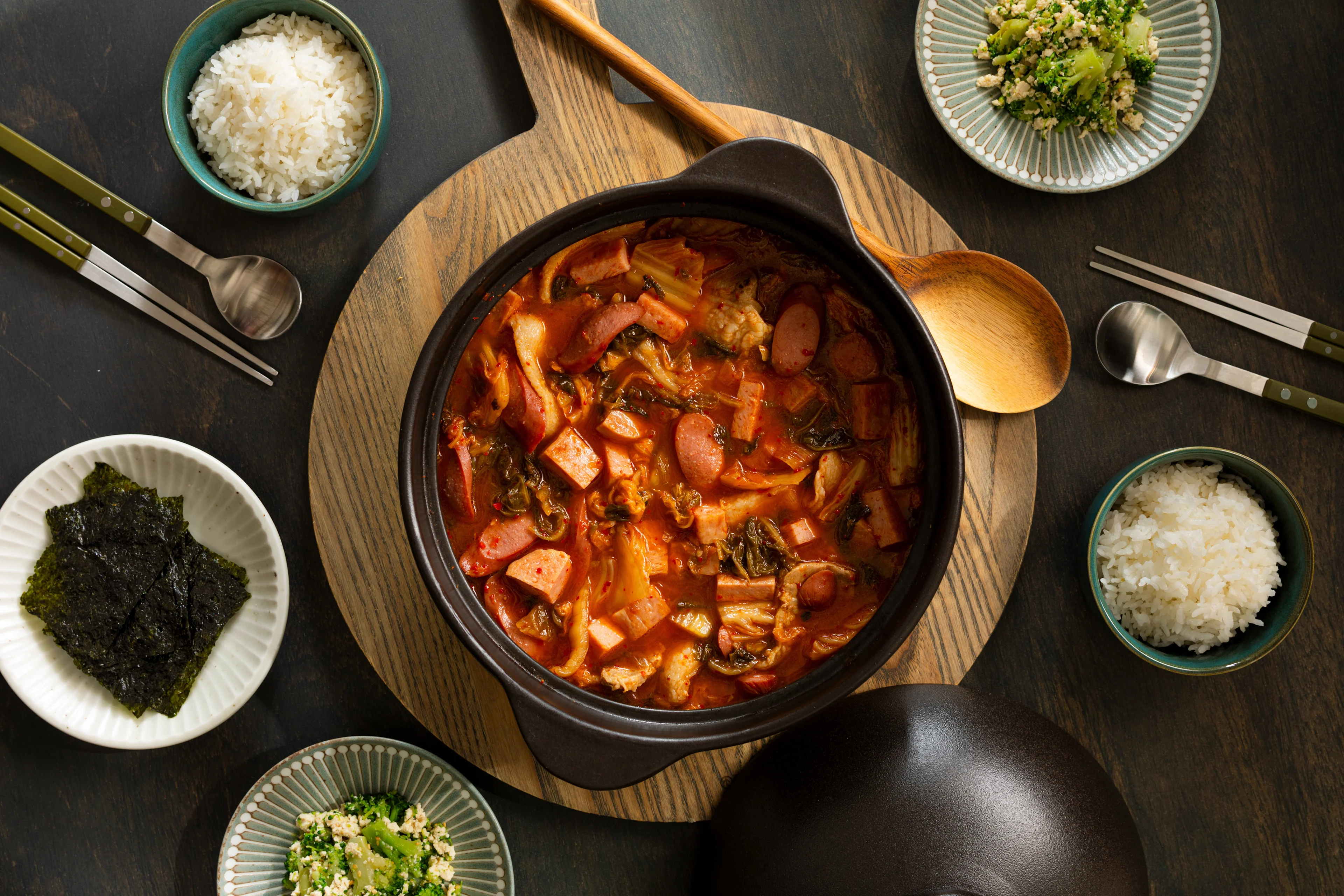 Korean stew with sausage and kimchi in black pot, surrounded by rice bowls, seaweed, and side dishes on dark table.