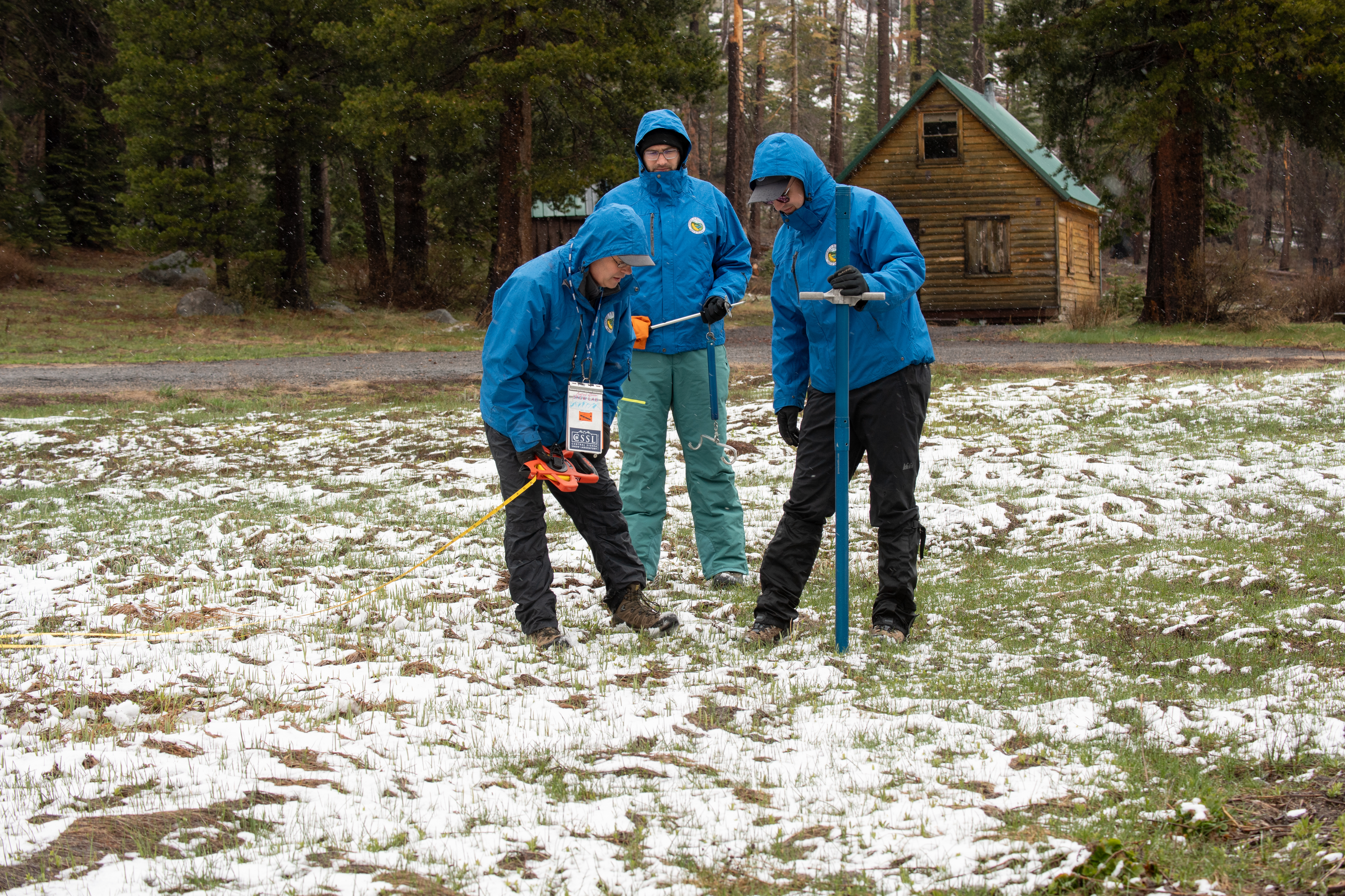 Three men in puffy blue coats who work for the state measure the meager snowpack in the Sierra Nevada Mountains with tube-like instruments. They are surrounded by some snow but mostly grass.