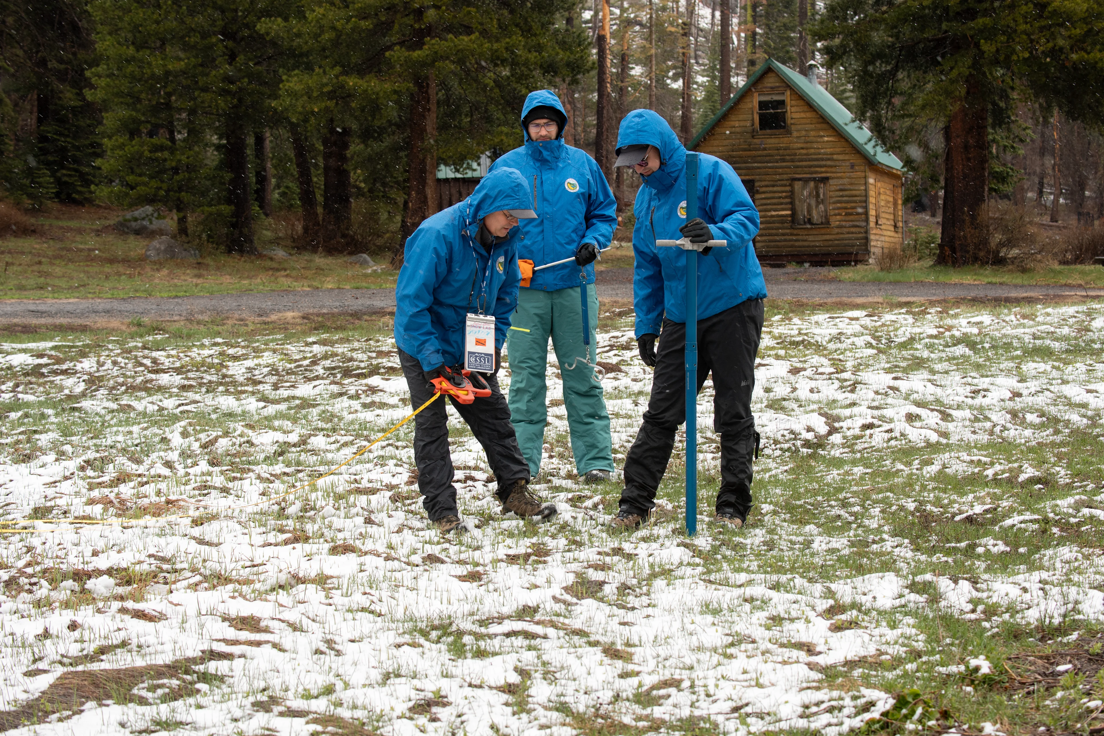 Three men in puffy blue coats who work for the state measure the meager snowpack in the Sierra Nevada Mountains with tube-like instruments. They are surrounded by some snow but mostly grass.