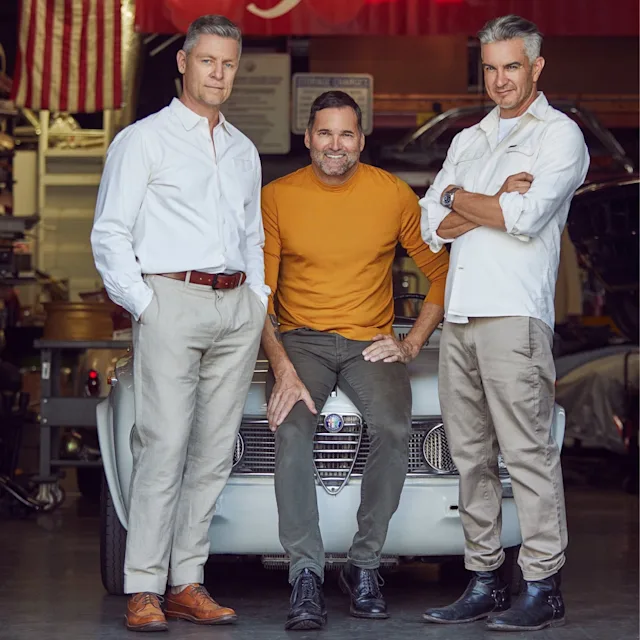Three men posing with vintage Alfa Romeo car in auto shop with American flag and red Alfa Romeo banner visible.