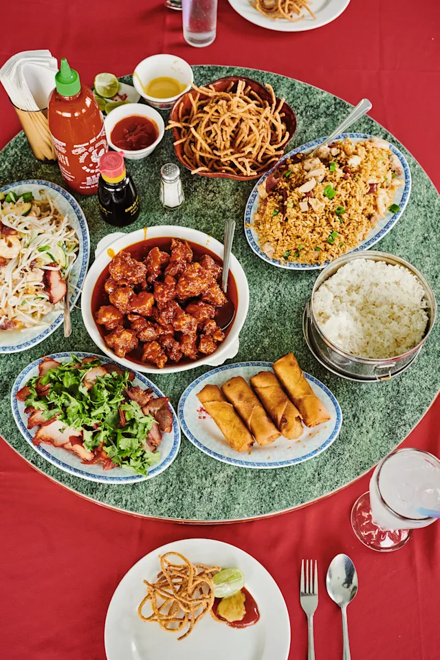 A Chinese restaurant table with fried rice, spring rolls, saucy chicken, roast meat, noodles, and white rice on a green lazy Susan.
