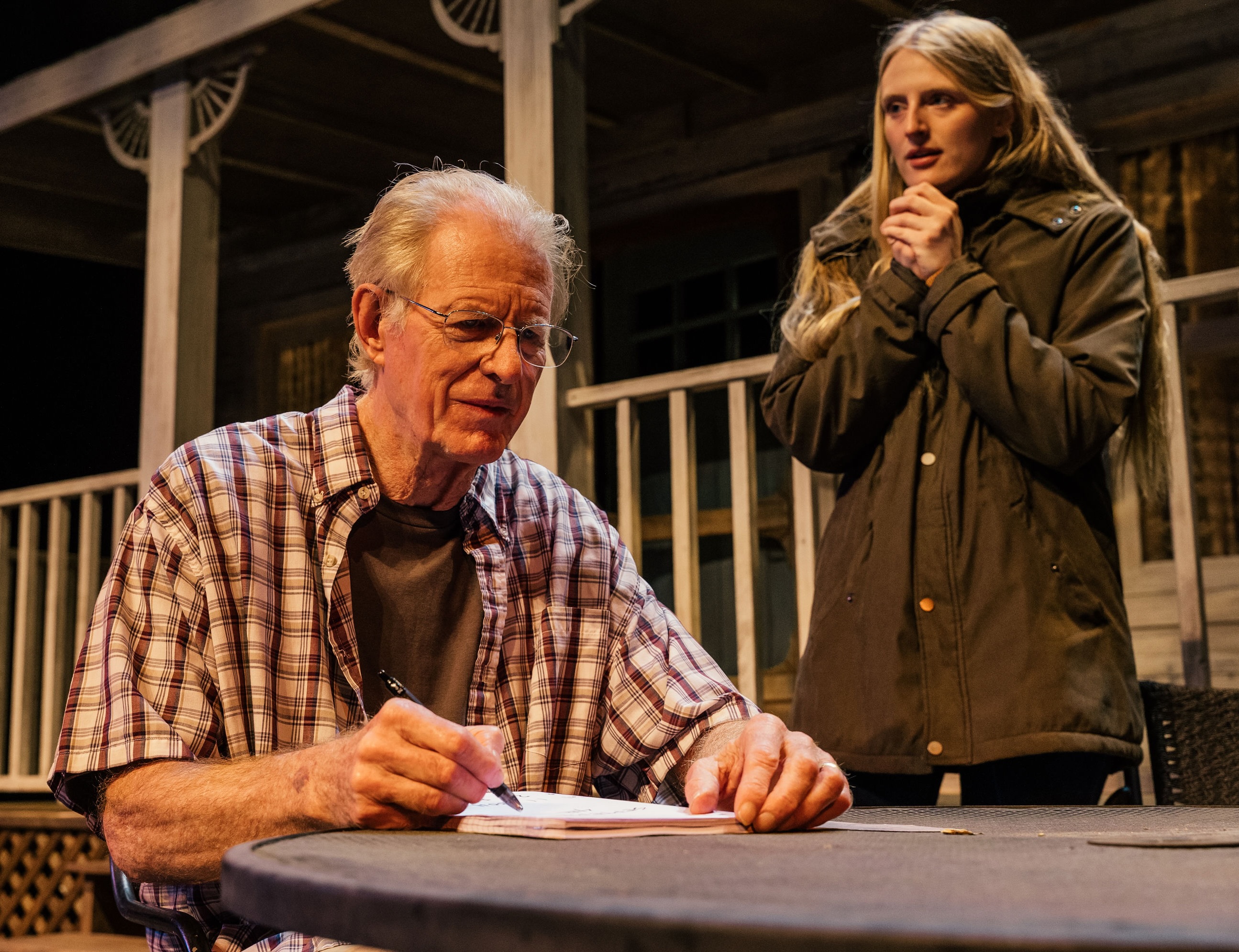 The actor Ed Begley Jr. sits at a table on a theater stage writing something, while his daughter and co-star Hayden Belgely looks on