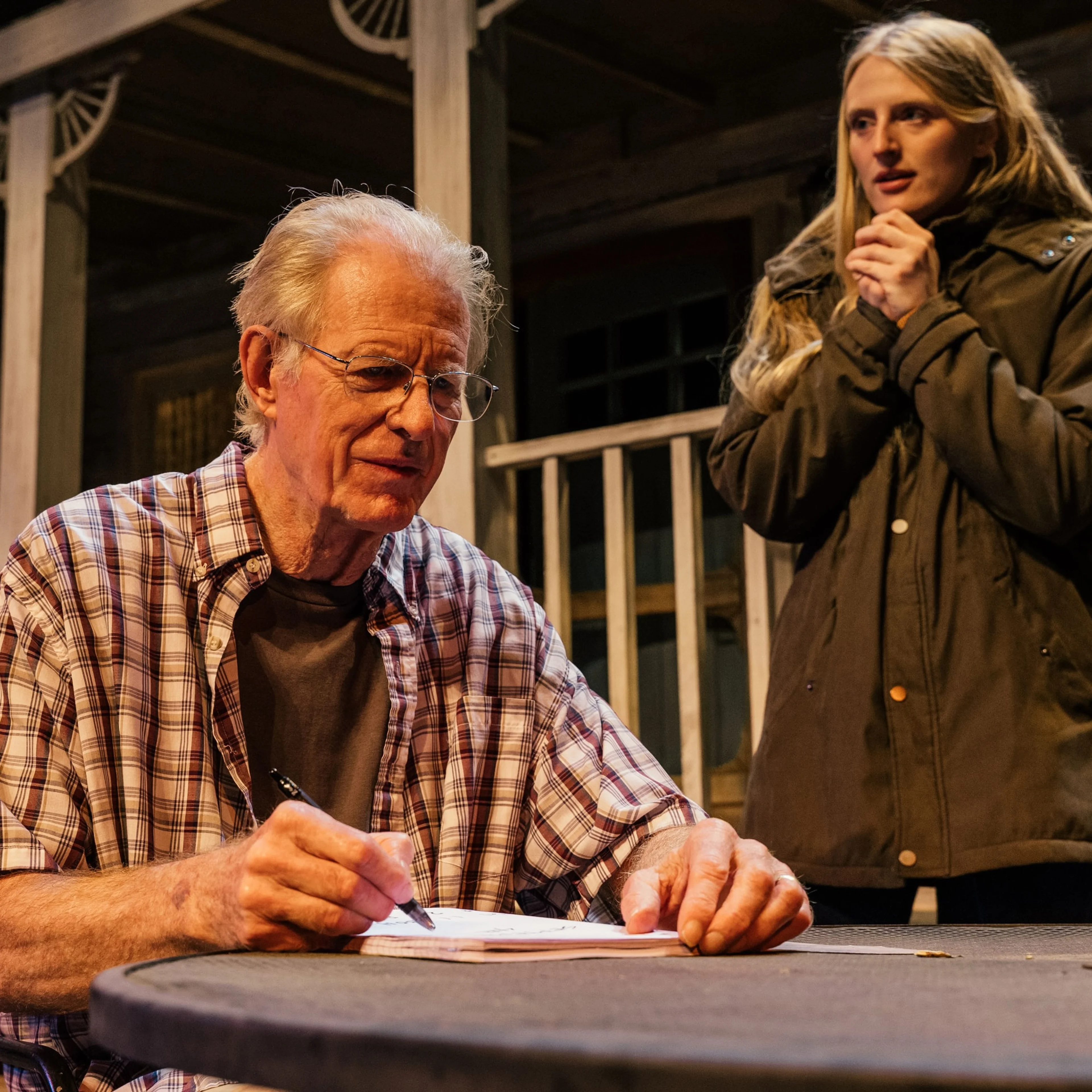 The actor Ed Begley Jr. sits at a table on a theater stage writing something, while his daughter and co-star Hayden Belgely looks on