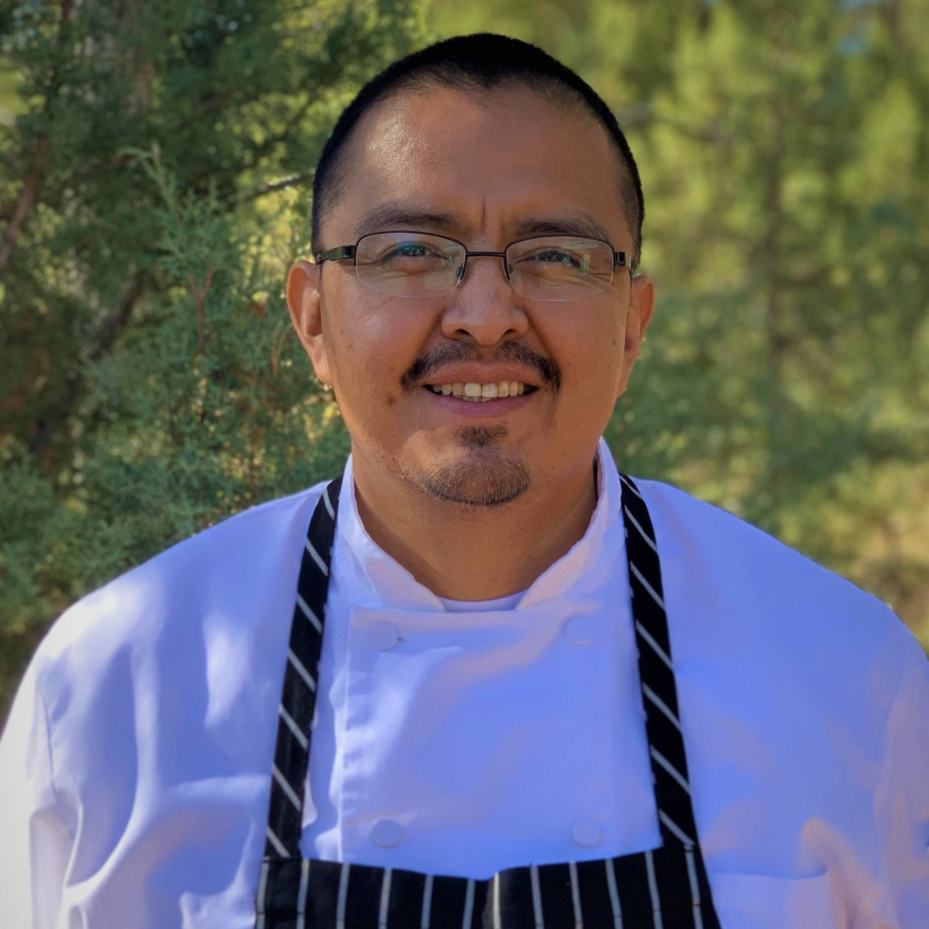 A smiling male chef wearing a white coat and black striped apron, with glasses, outdoors with green trees behind him.