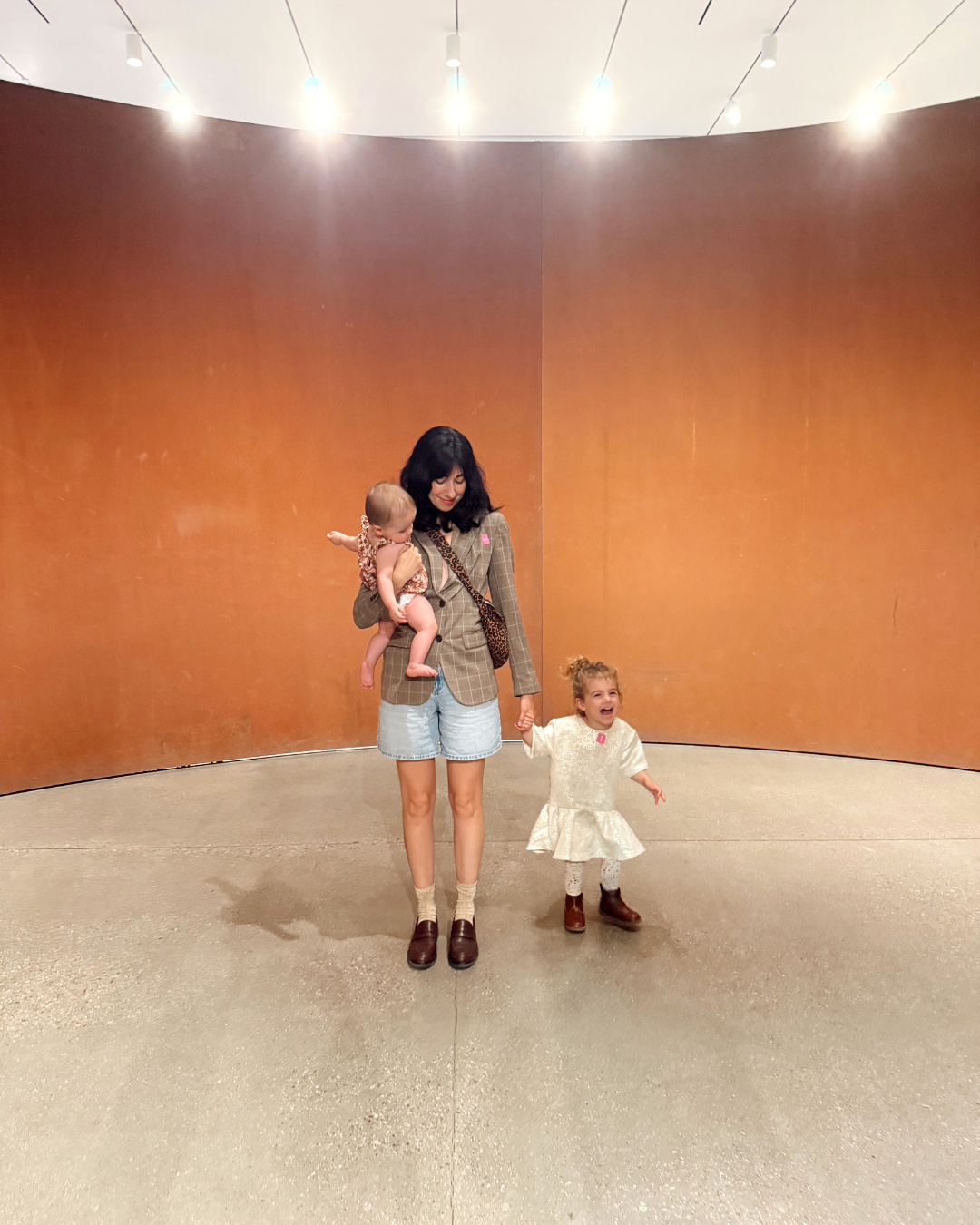A woman holds a baby while a toddler in a white dress laughs beside her in a modern art gallery with terracotta walls.