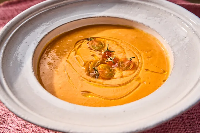 A white ceramic bowl of creamy orange tomato soup topped with cherry tomatoes, herbs, and a drizzle of olive oil.