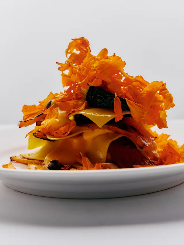A white plate with a stacked dish of yellow pasta or squash ribbons topped with vibrant orange dried flower petals.