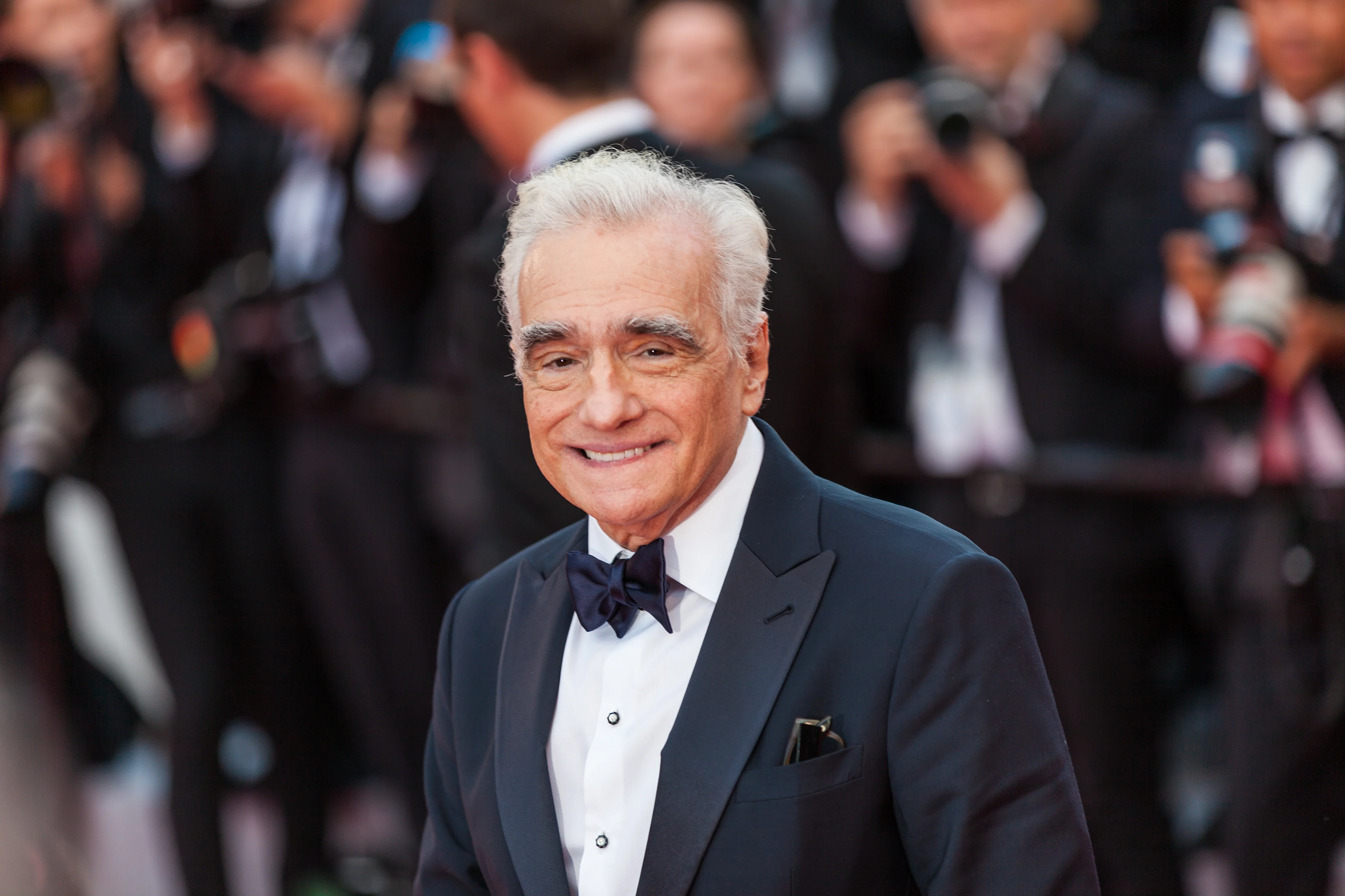 Martin Scorsese with white hair in a black tuxedo smiling at a formal event with blurred crowd in background.
