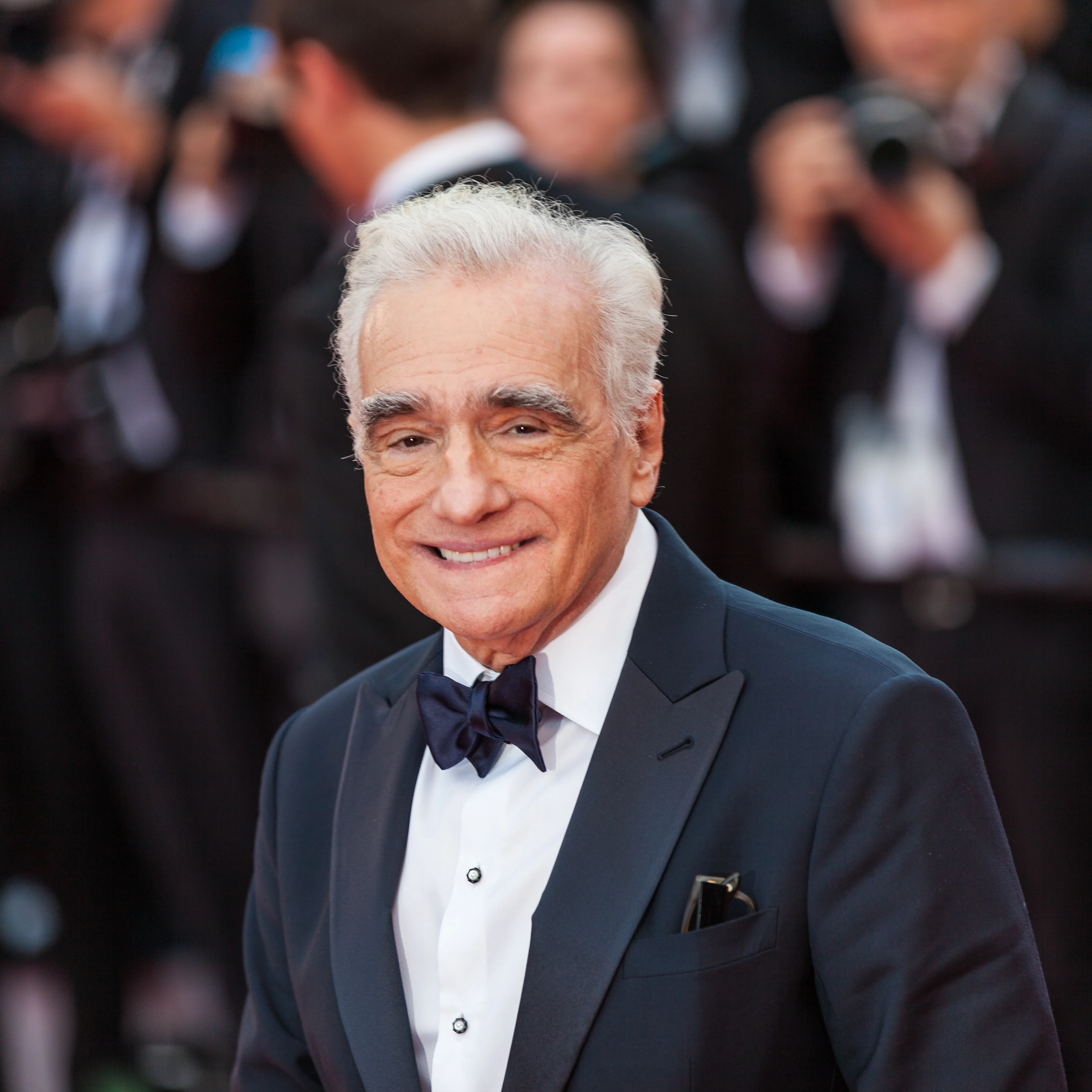 Martin Scorsese with white hair in a black tuxedo smiling at a formal event with blurred crowd in background.