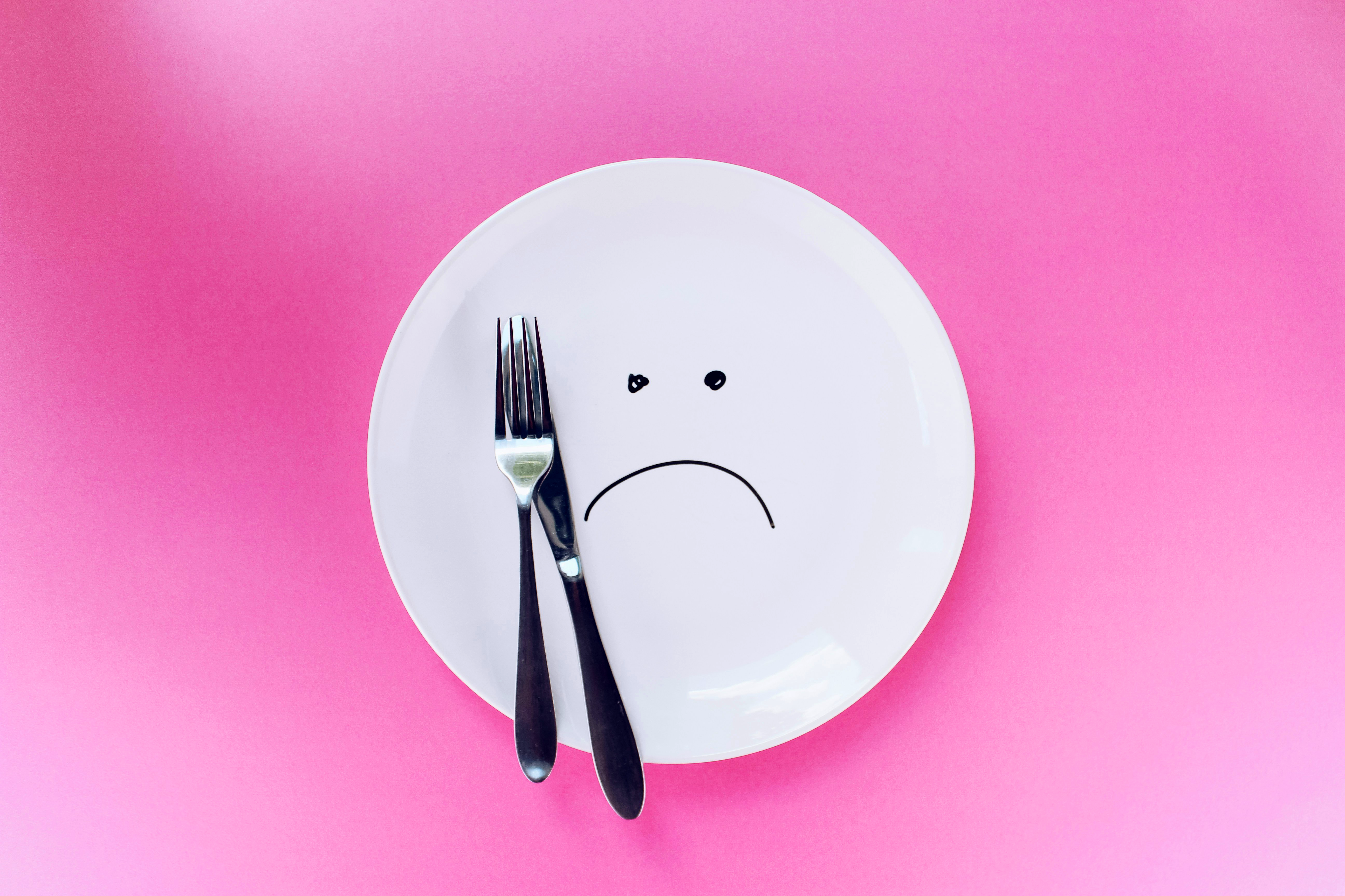 A white plate with a drawn sad face sits on a pink background, with a silver fork and knife resting on it.